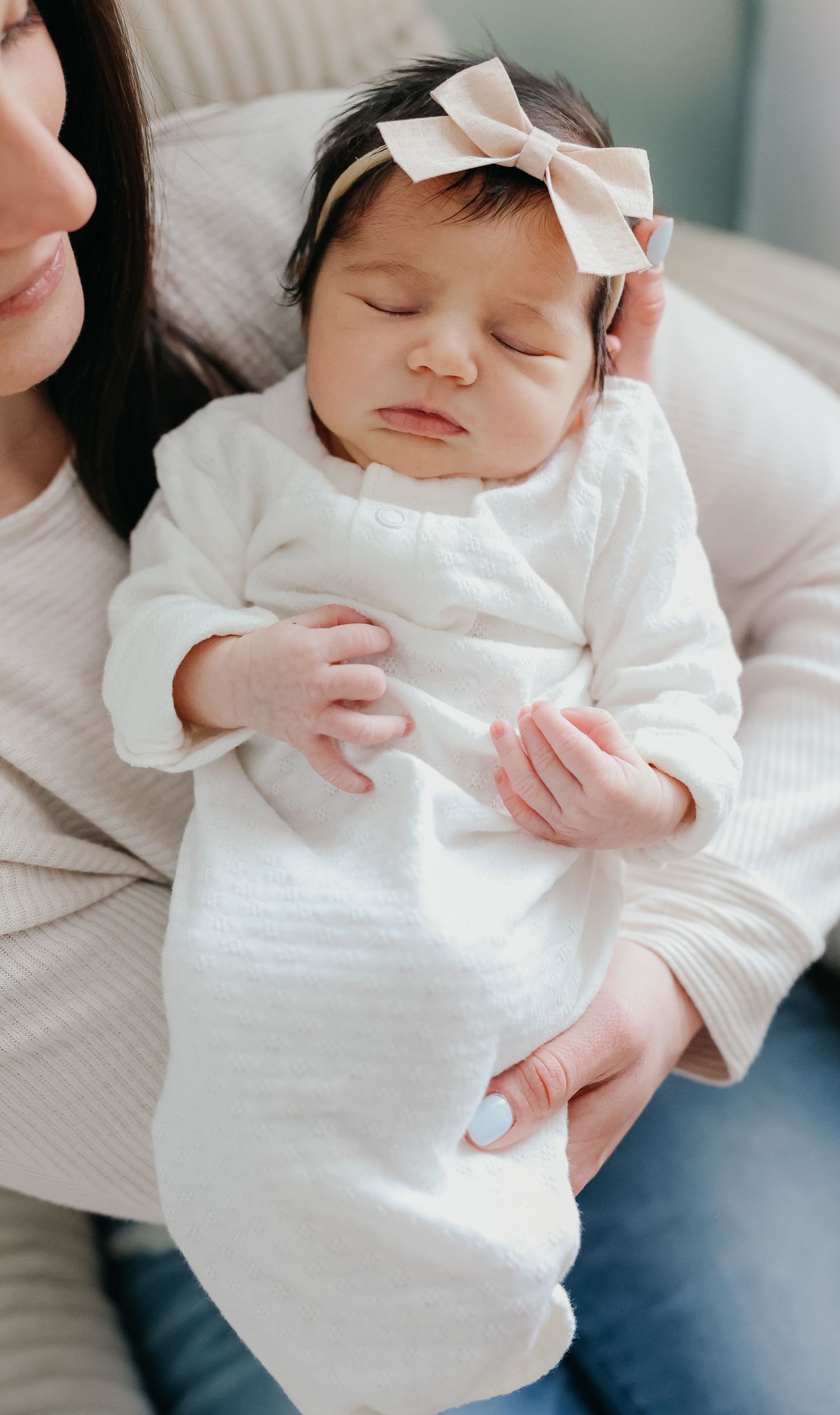 Sleeping baby in white outfit with bow headband resting on caregiver's lap.