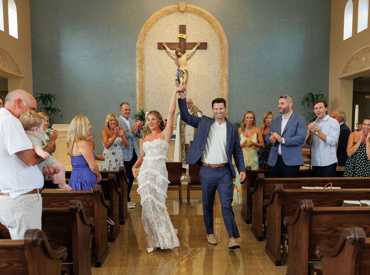 Joyful newlyweds celebrate after church wedding ceremony, guests applauding in chapel with crucifix backdrop.