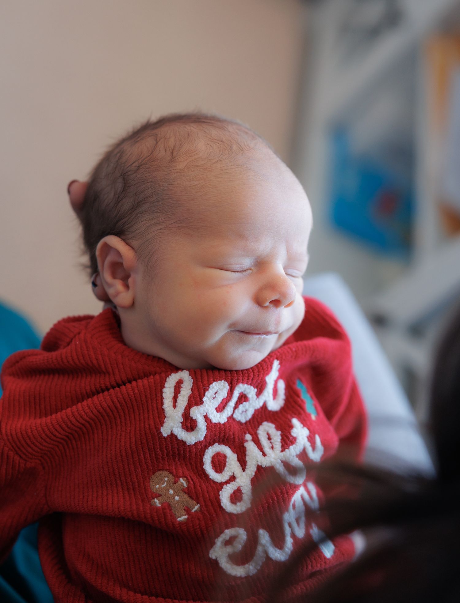 A sleeping newborn baby wearing a red knit sweater with white text embroidered on it.