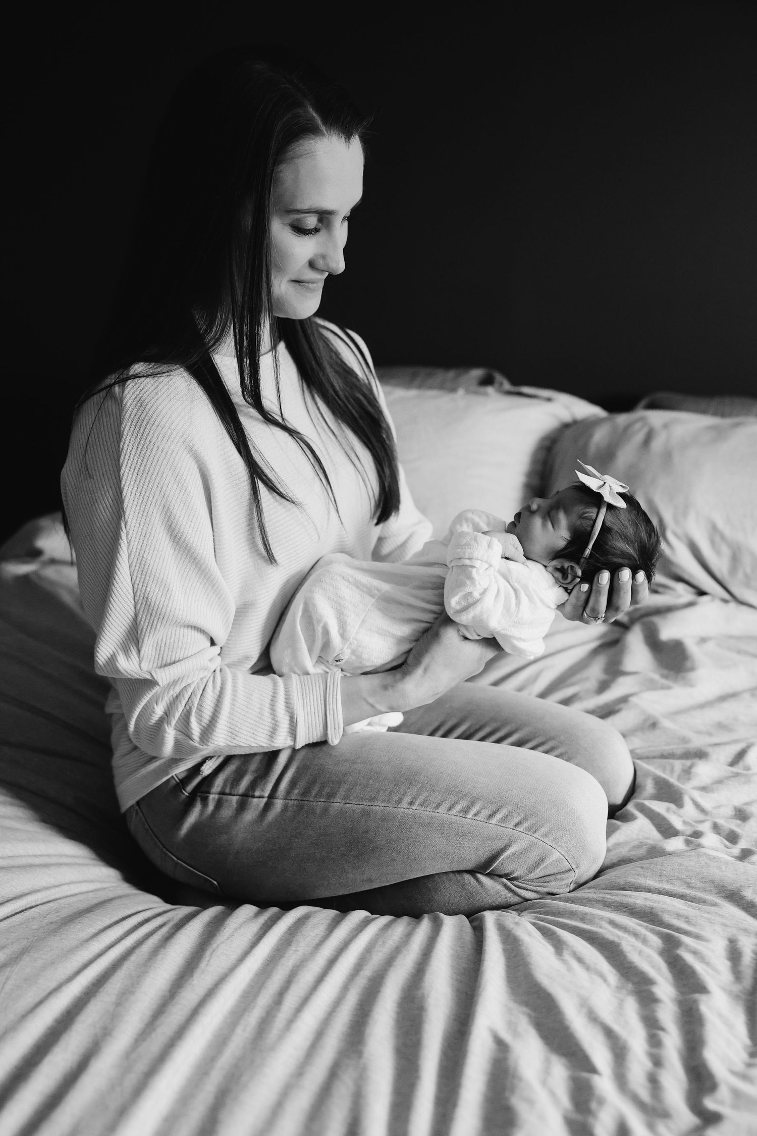 Person in a white sweater sitting on a bed holding a sleeping newborn in black and white photography.