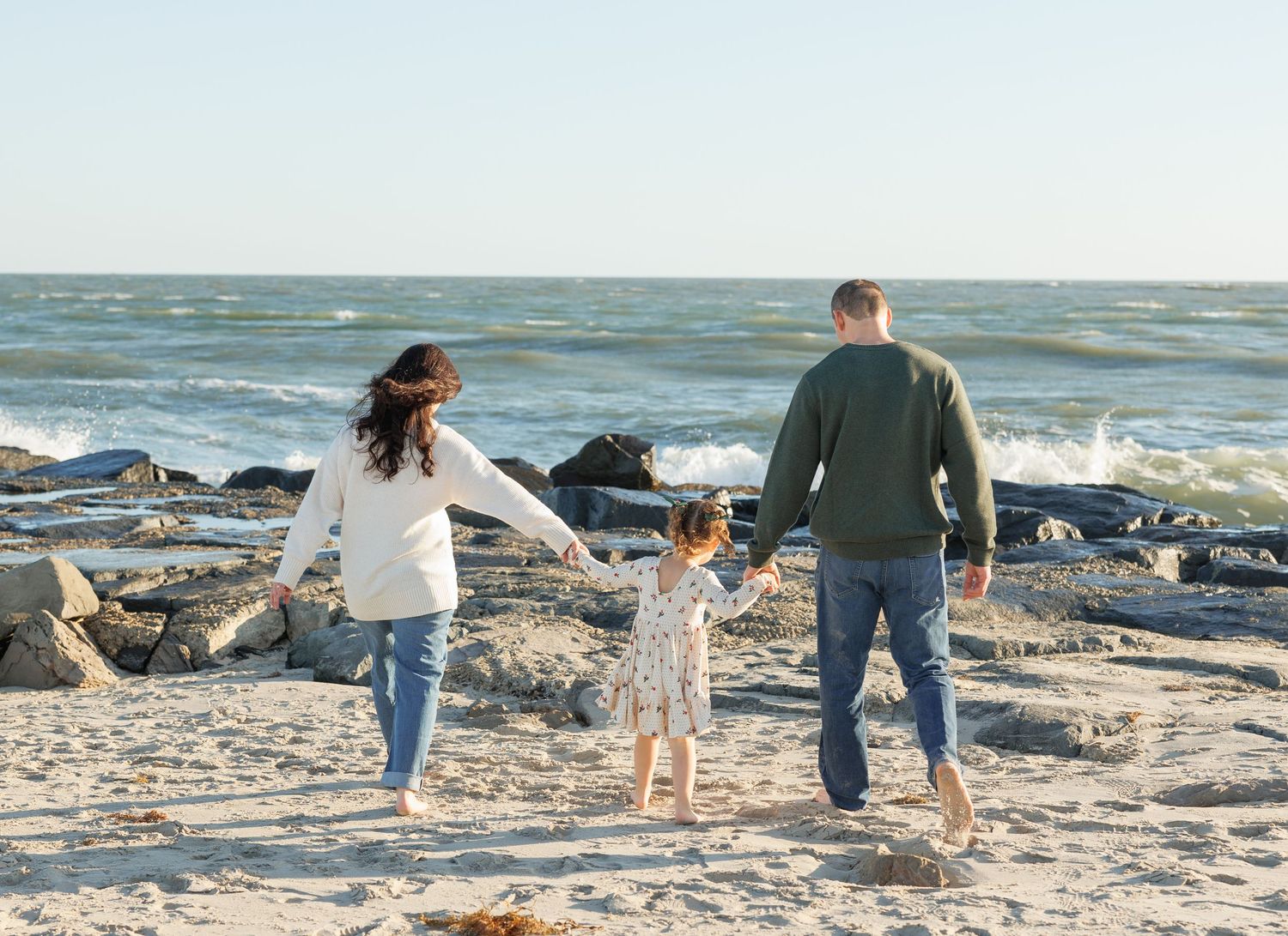 Family holding hands while walking together on a rocky beach with ocean waves in the background.