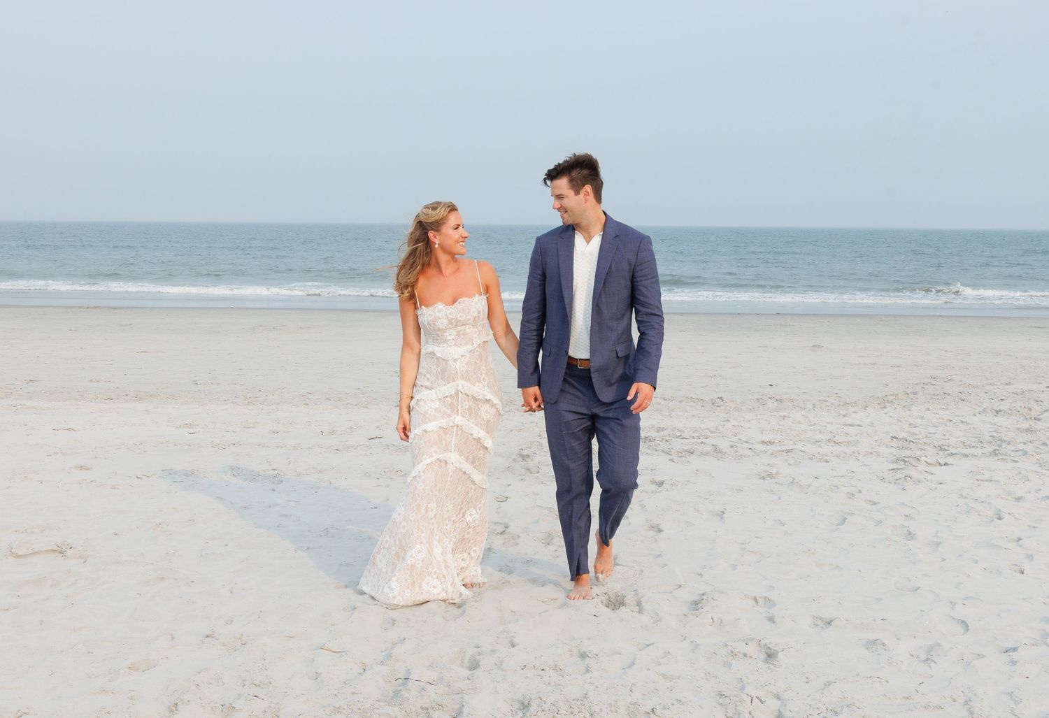 Bride and groom walk hand-in-hand on a sandy beach, she in a lace gown, he in a blue suit.