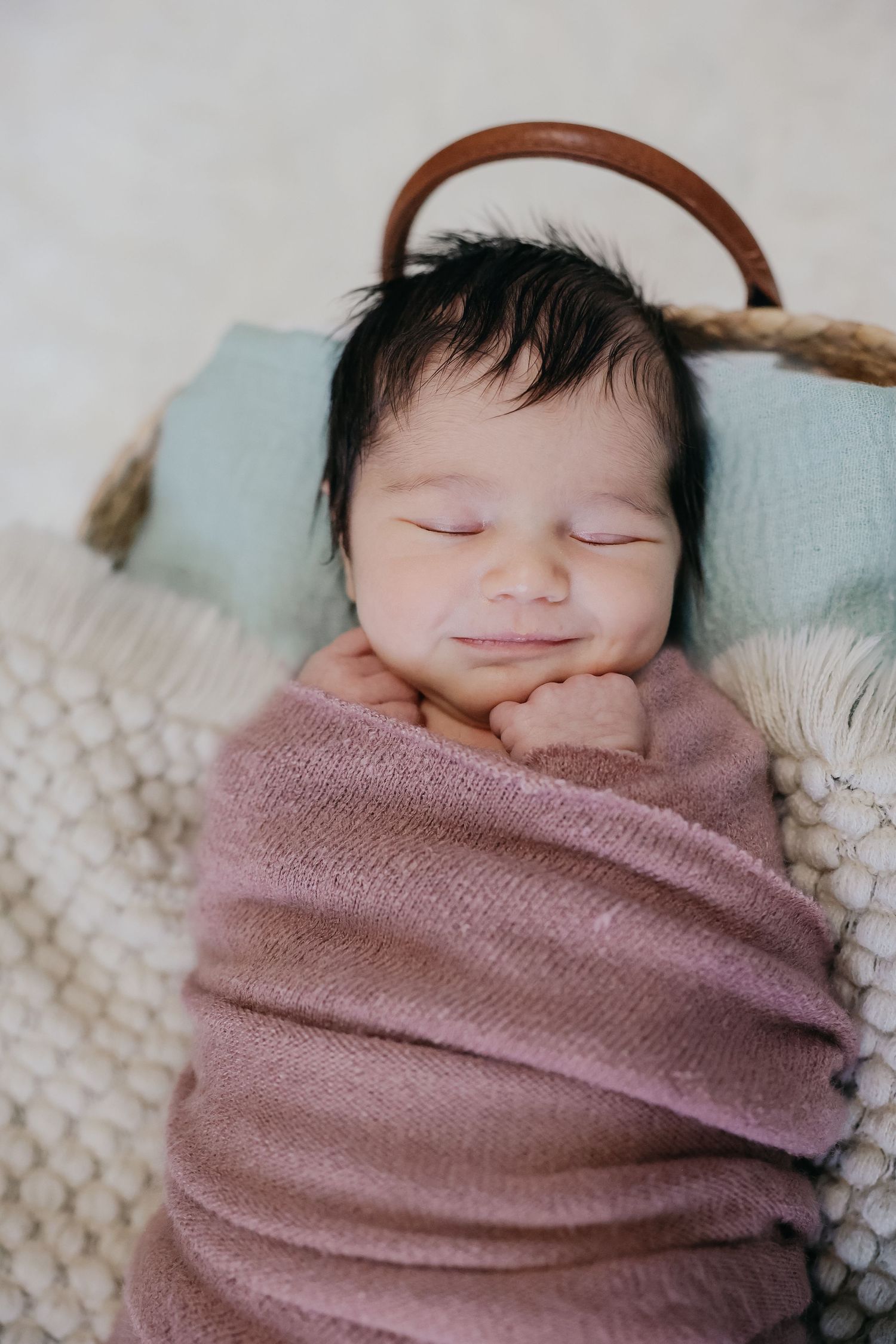 Sleeping newborn wrapped in pink swaddle with knit headband on textured blanket.