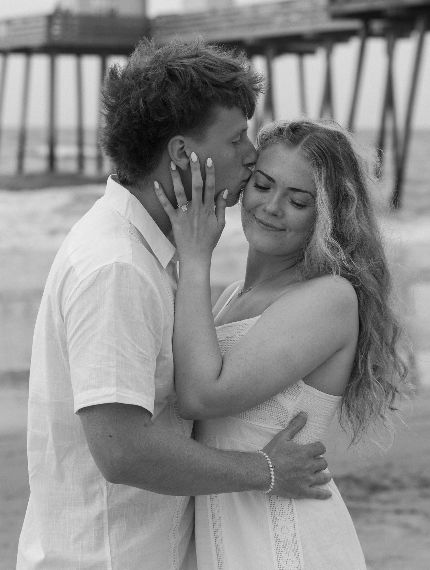 Black and white beach photo of a couple embracing and kissing near a pier, woman showing engagement ring.