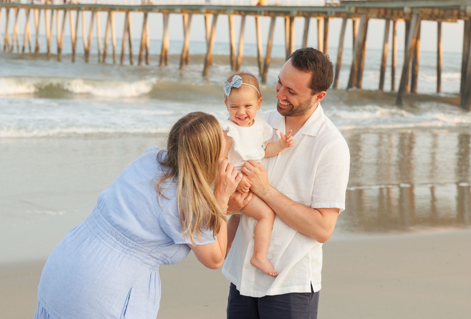 Family beach photo with mom leaning in to kiss baby held by smiling dad near ocean pier.