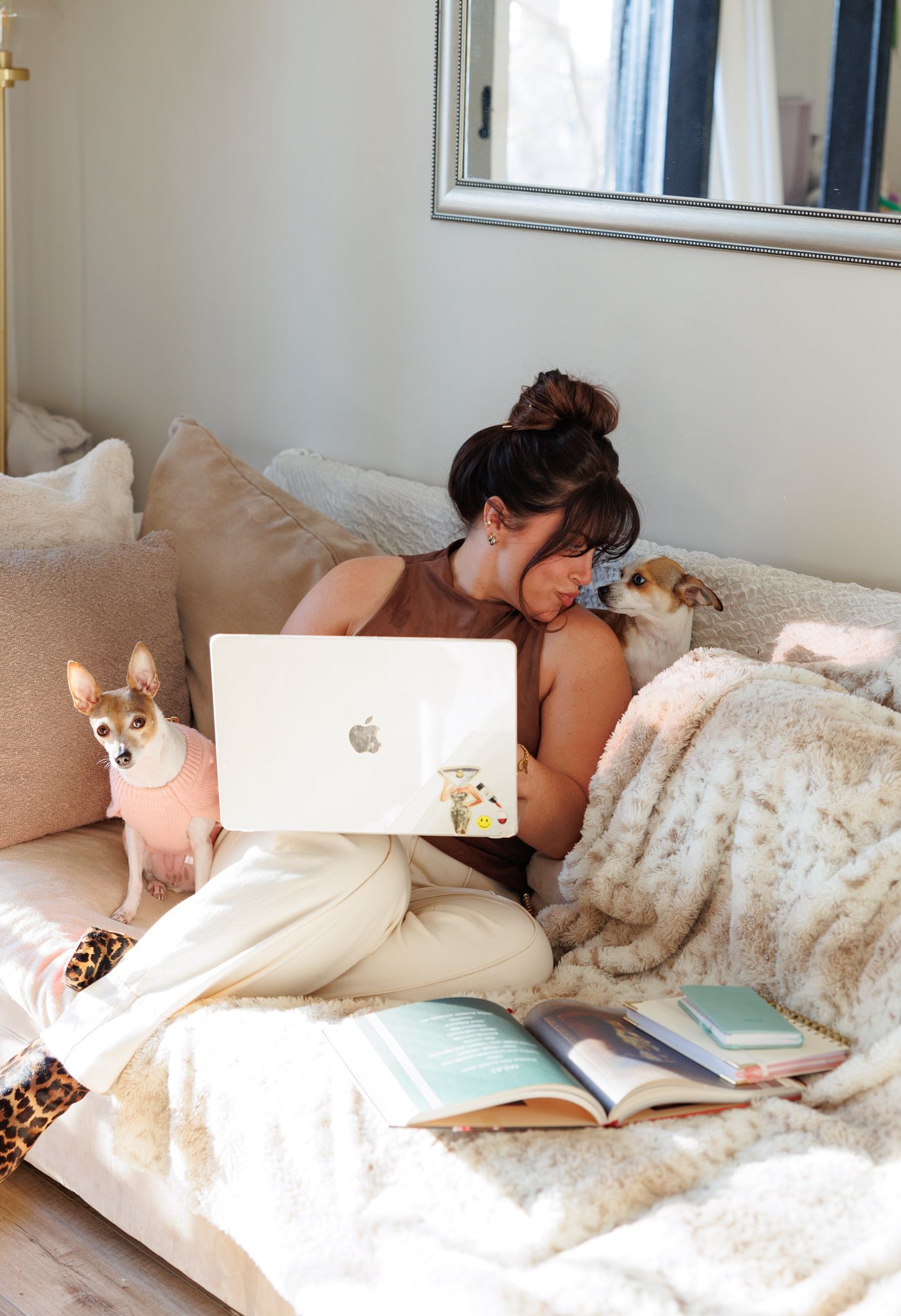 Woman sitting on couch with MacBook and two dogs, surrounded by books and cozy blankets in a bright living space.