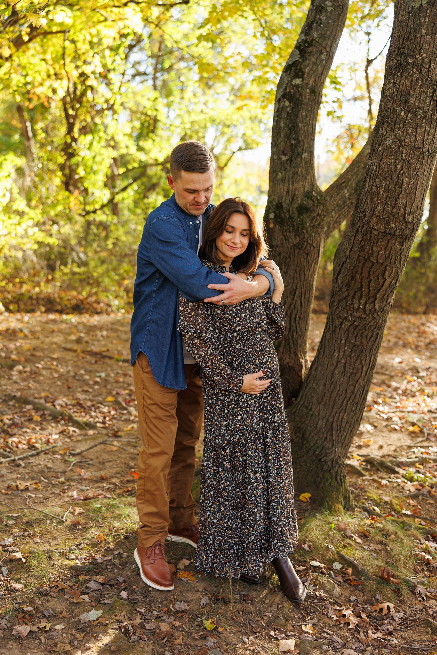 A couple shares a tender moment among autumn trees with fallen leaves scattered on the ground.