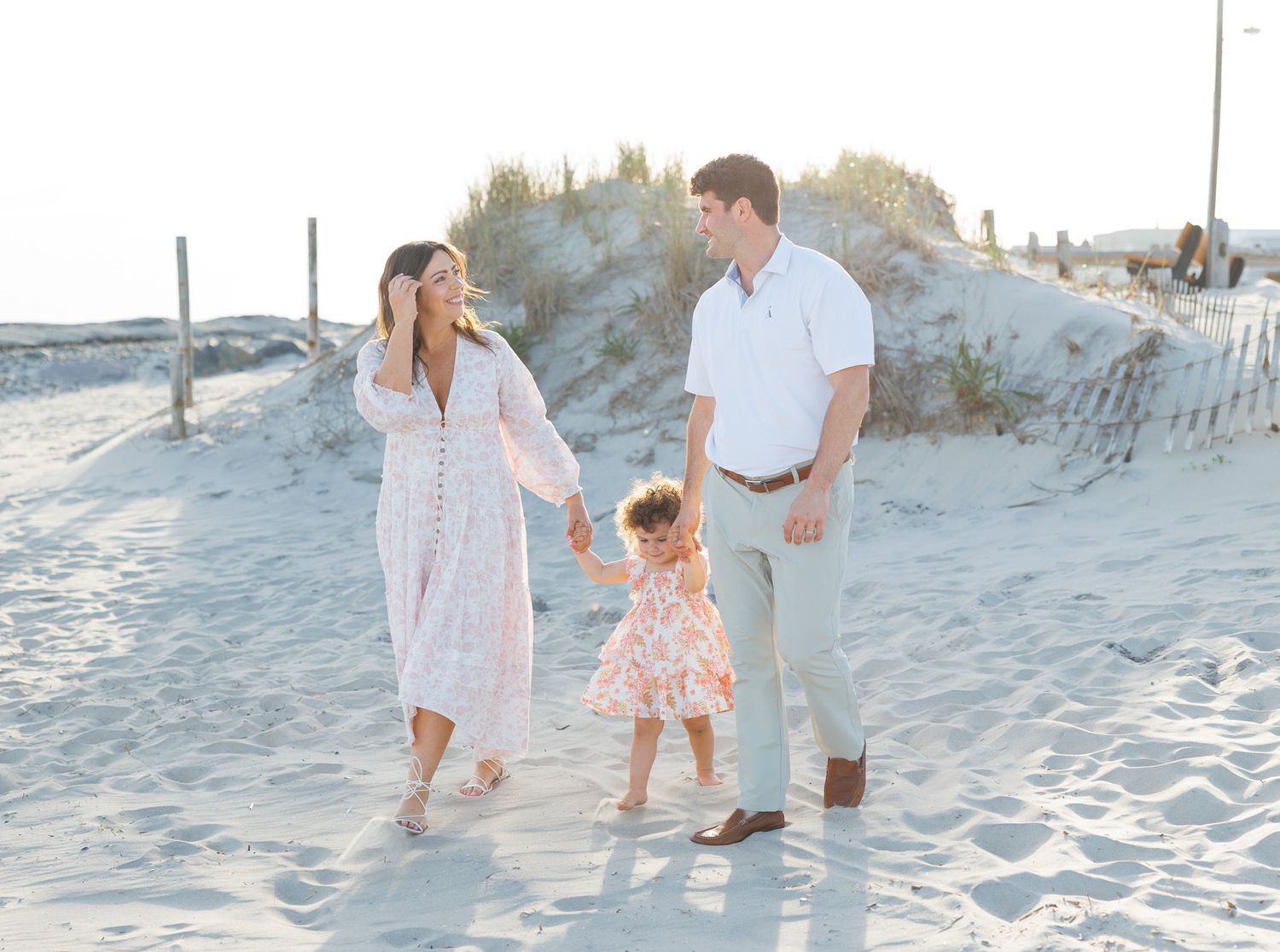 A family wearing light colored clothing walks together on a sandy beach at sunset with dunes in the background.