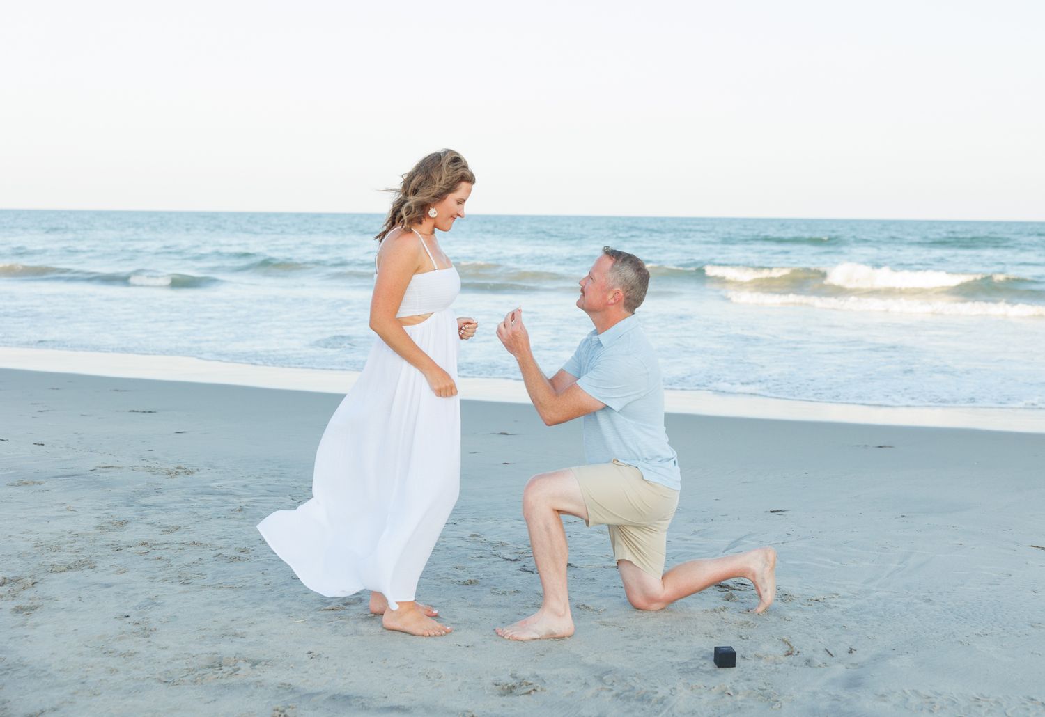 A couple in white clothing shares a romantic proposal moment on a sandy beach at sunset.