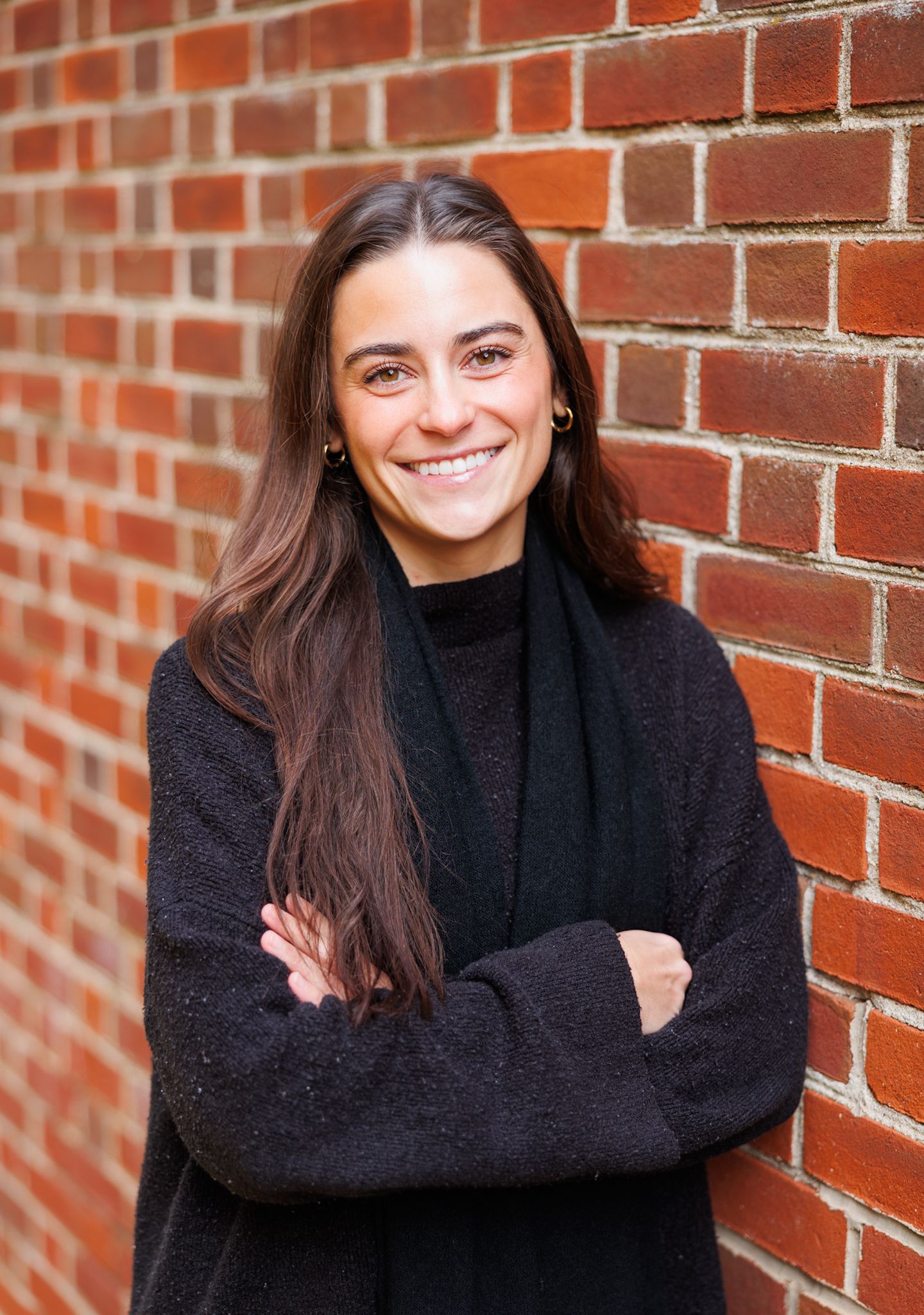 Smiling young woman with long brown hair in black outfit, arms crossed, leaning against a red brick wall.