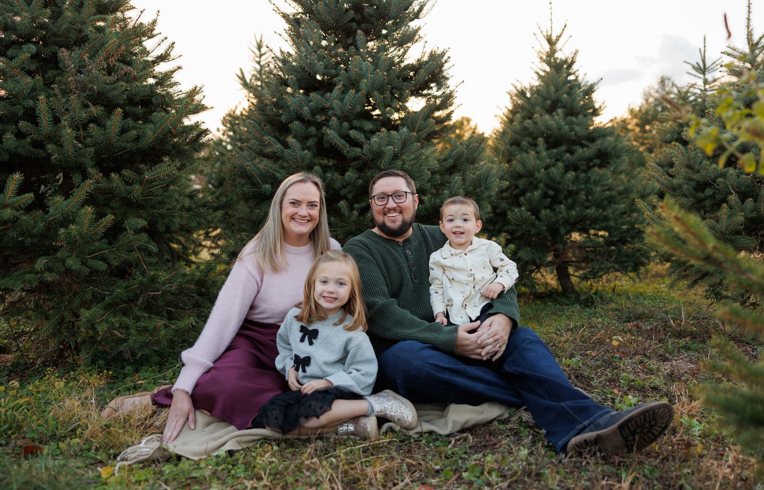 A family sits together on a blanket among evergreen trees during an outdoor autumn photo session.