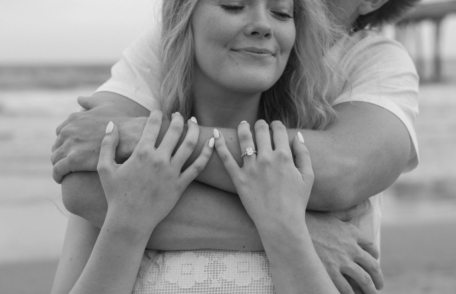 Black and white photo of a couple embracing on a beach, woman smiling with eyes closed, engagement ring visible.