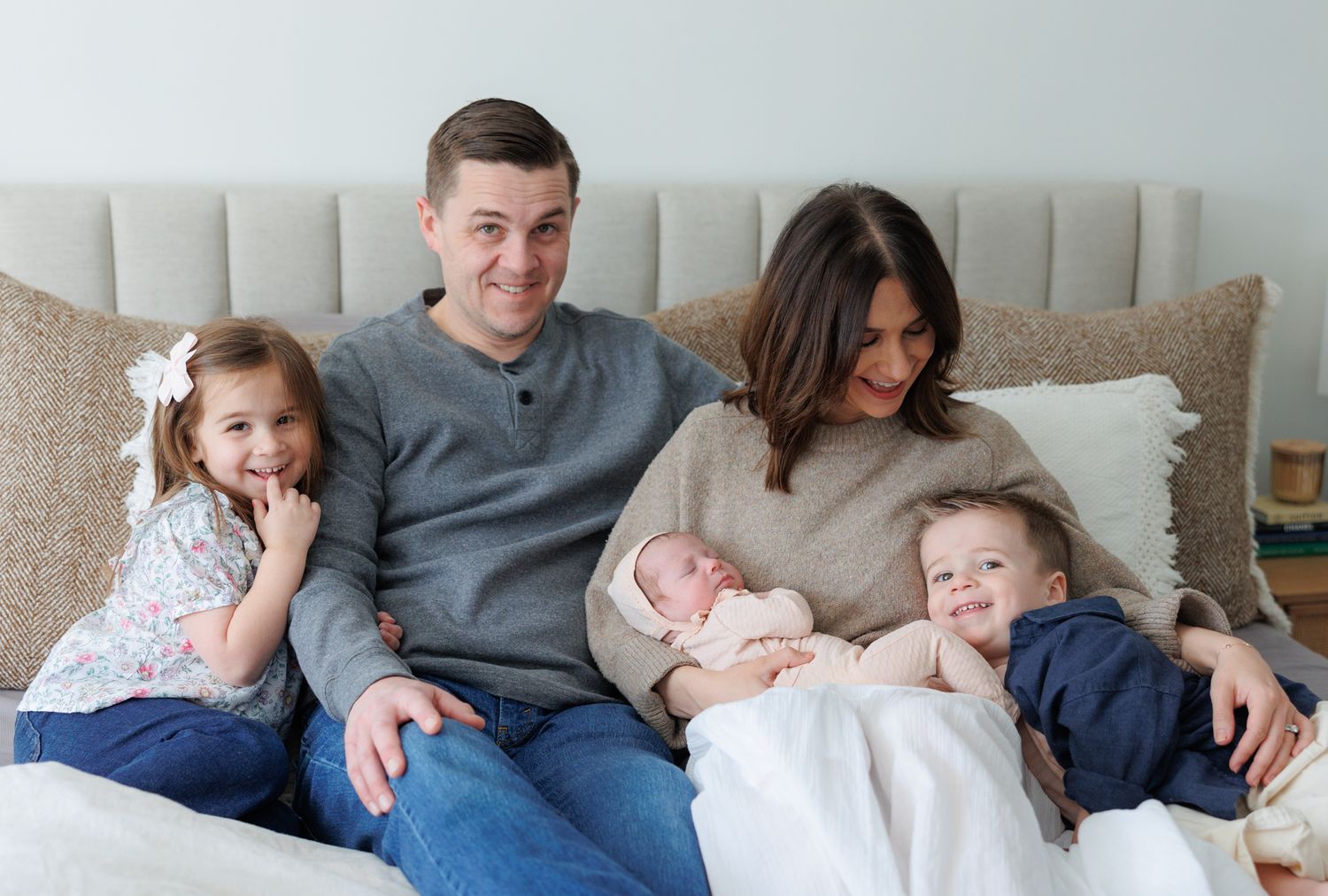 Happy family of five with newborn baby posing together on a white bed in a bright, cozy bedroom setting.