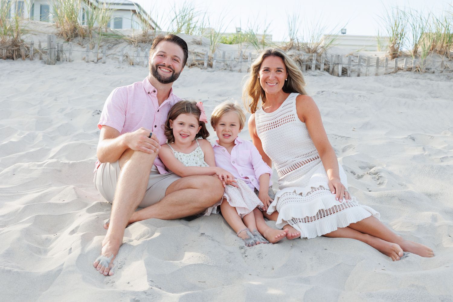 Family of four smiling together on a sandy beach, parents and two young children in coordinating pastel outfits.