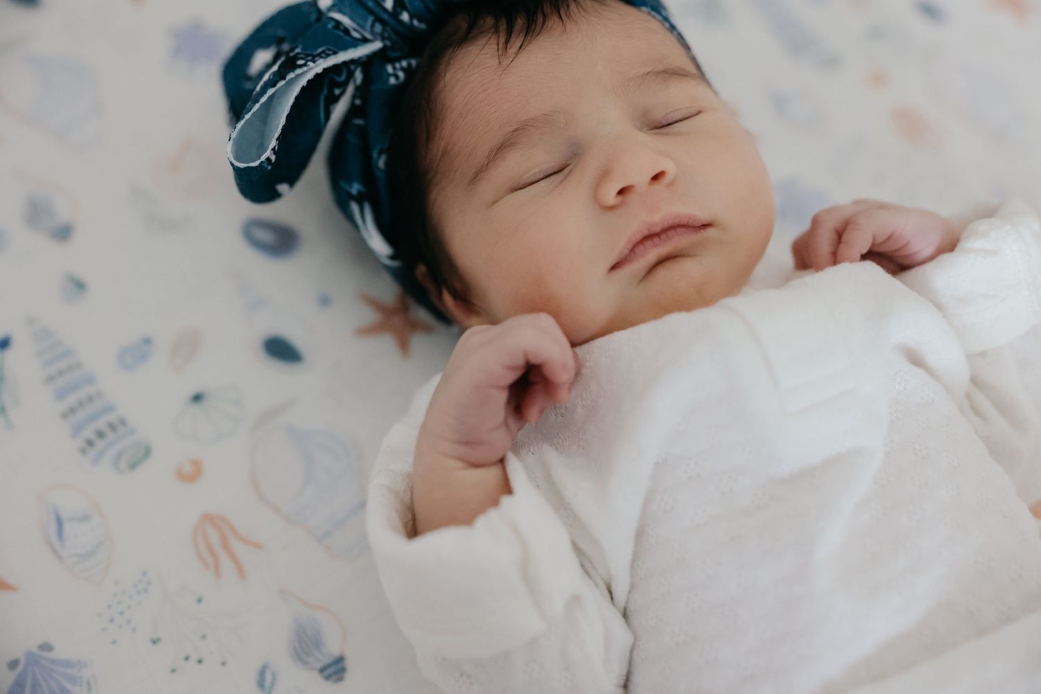 Close-up of peaceful sleeping baby wearing white outfit and blue bow on patterned fabric.