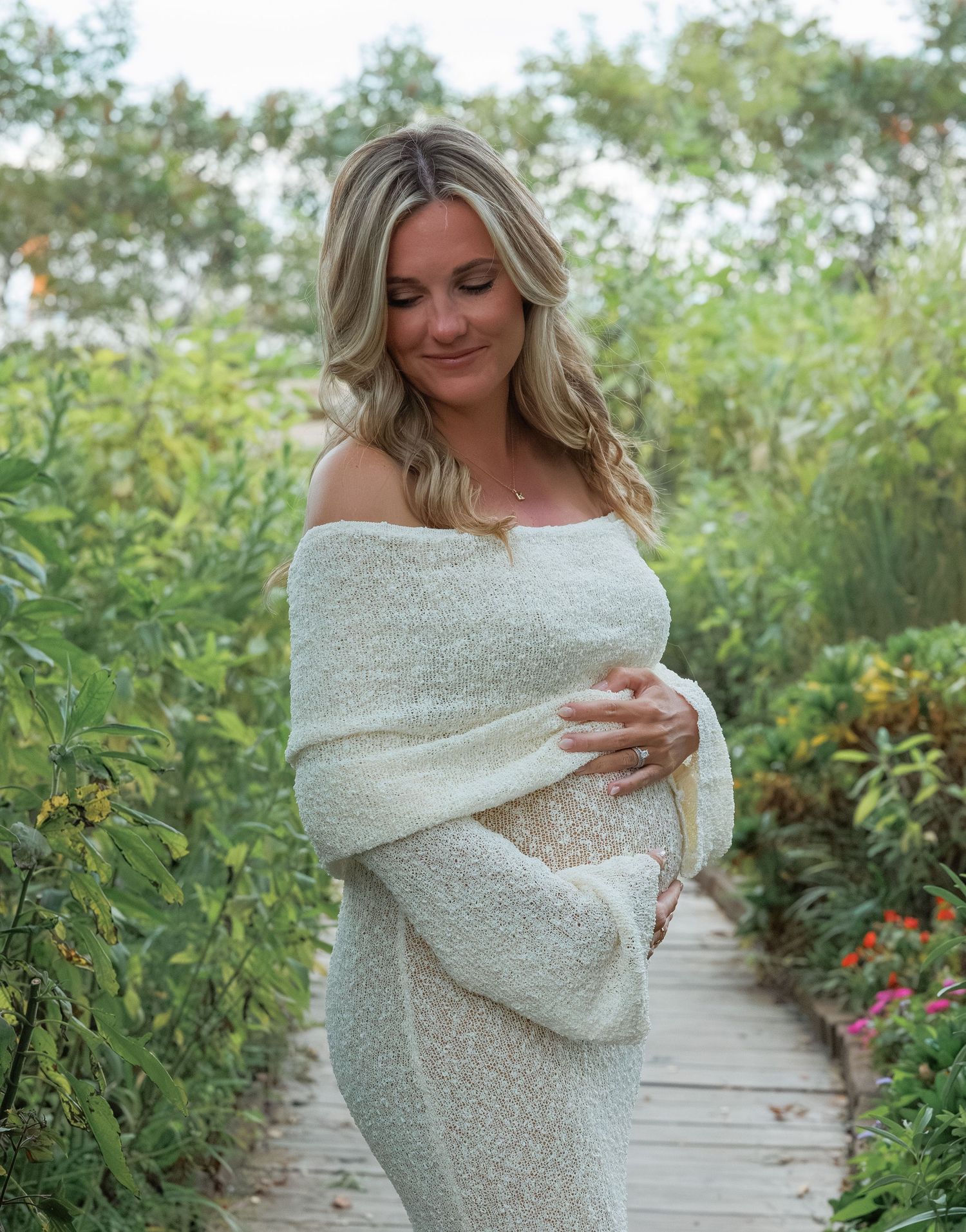 Person wearing a cozy off-shoulder sweater poses on wooden boardwalk surrounded by lush greenery and flowers.