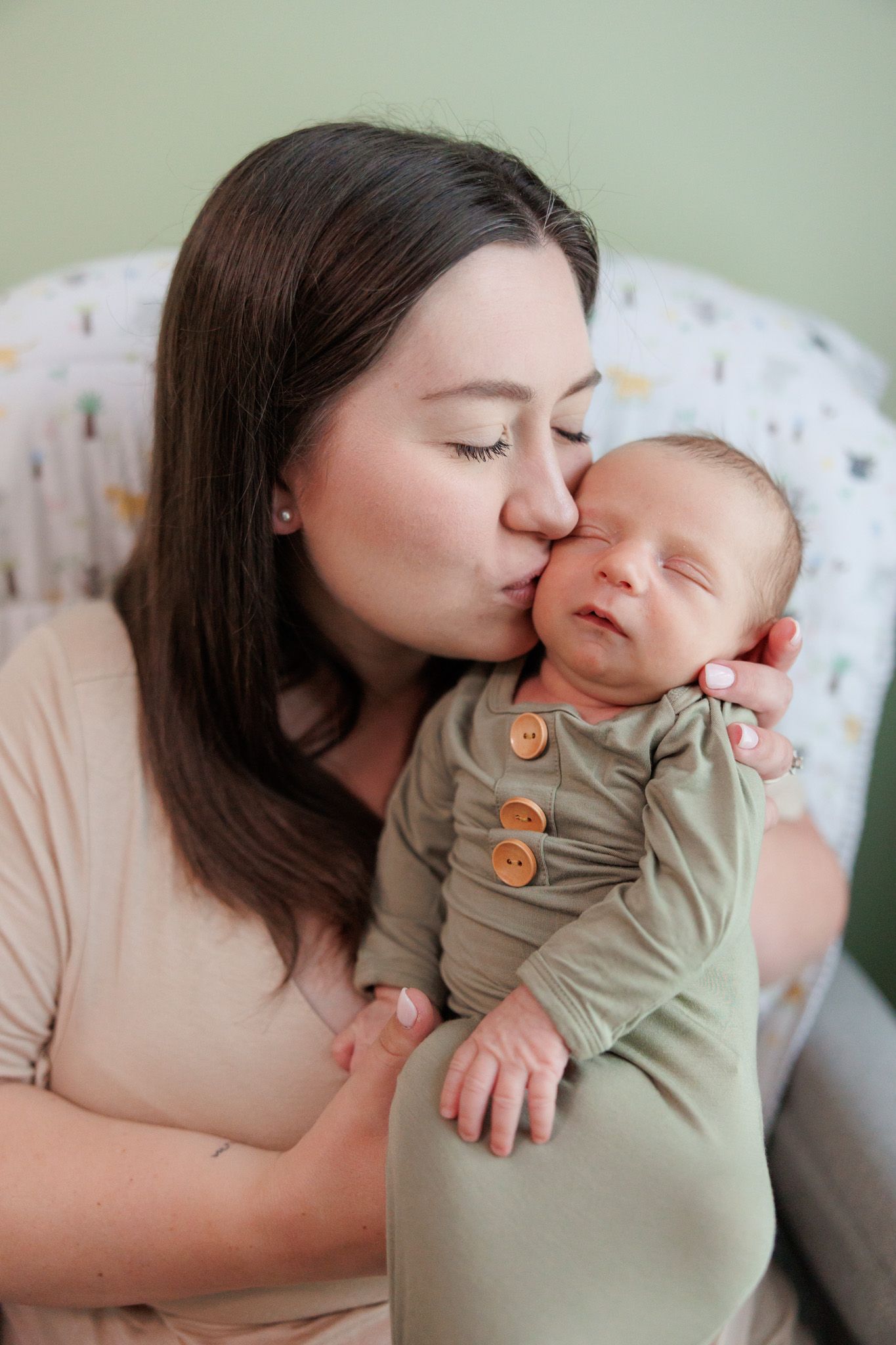 A tender moment between a mother and newborn baby as she gives a gentle kiss while holding them in a cozy chair.
