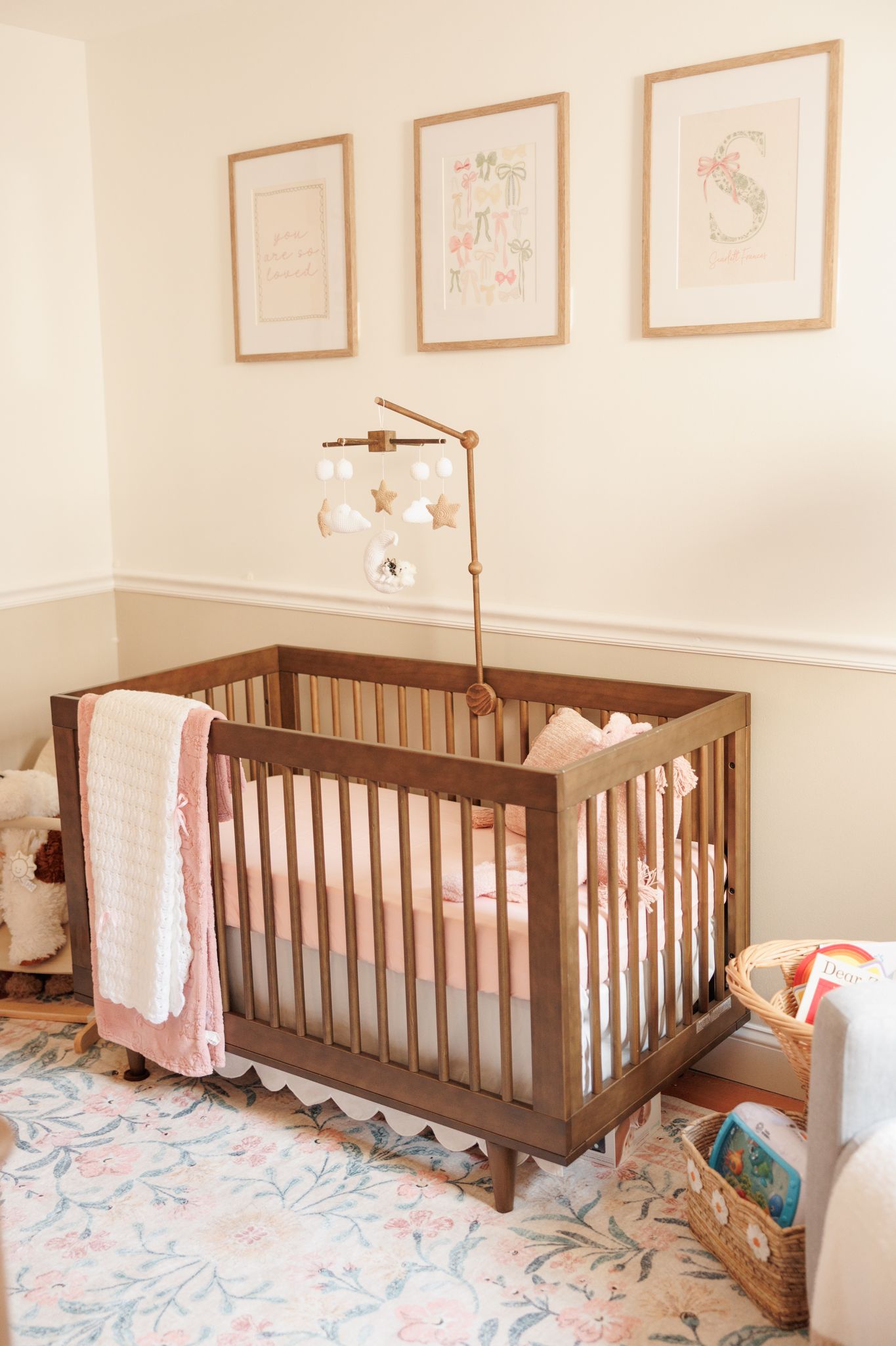 Wooden baby crib with pink blanket, mobile, floral rug, and three framed prints in a soft nursery.
