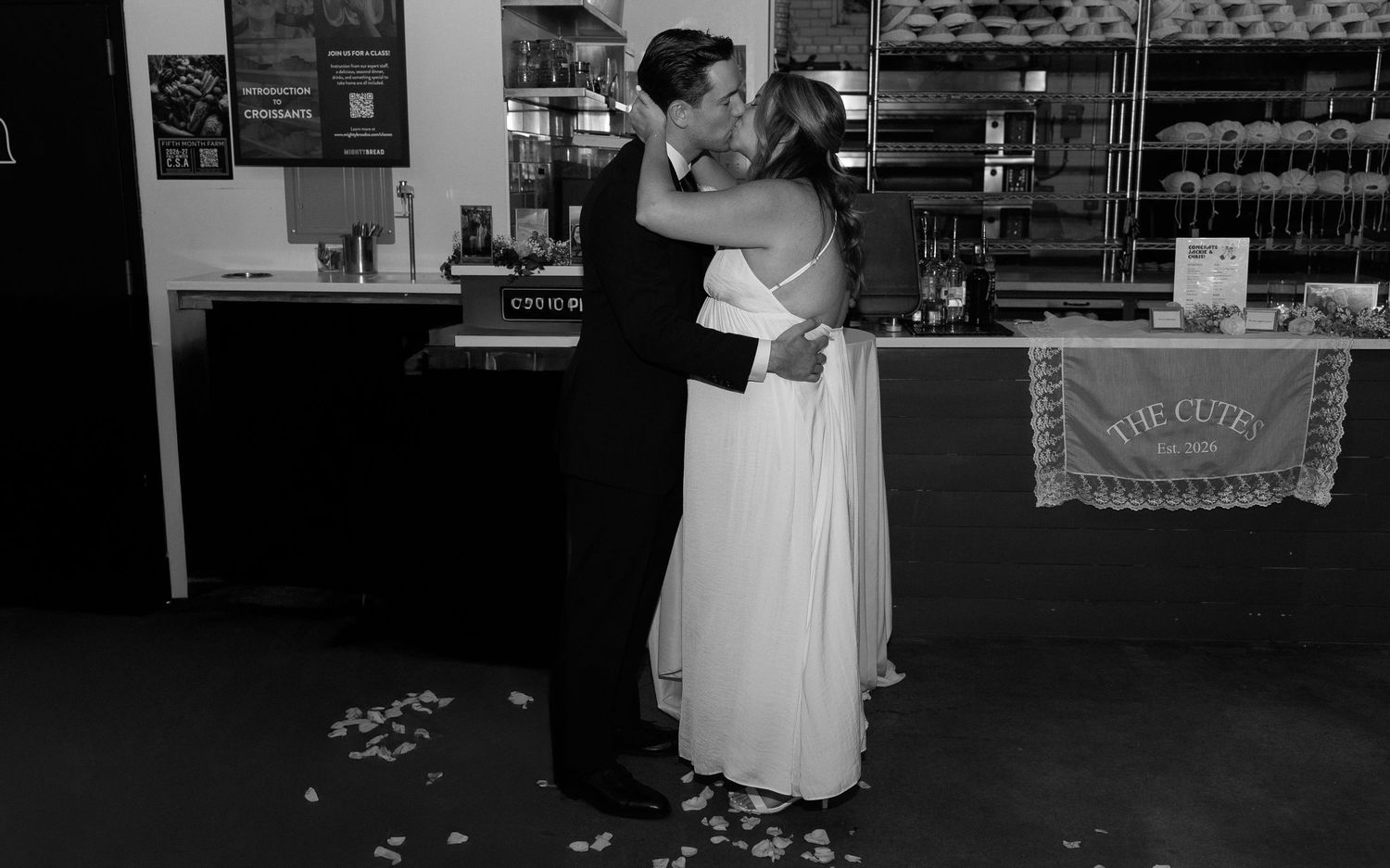 Black and white photo of a couple sharing a first dance kiss in wedding attire inside a cozy bakery cafe.