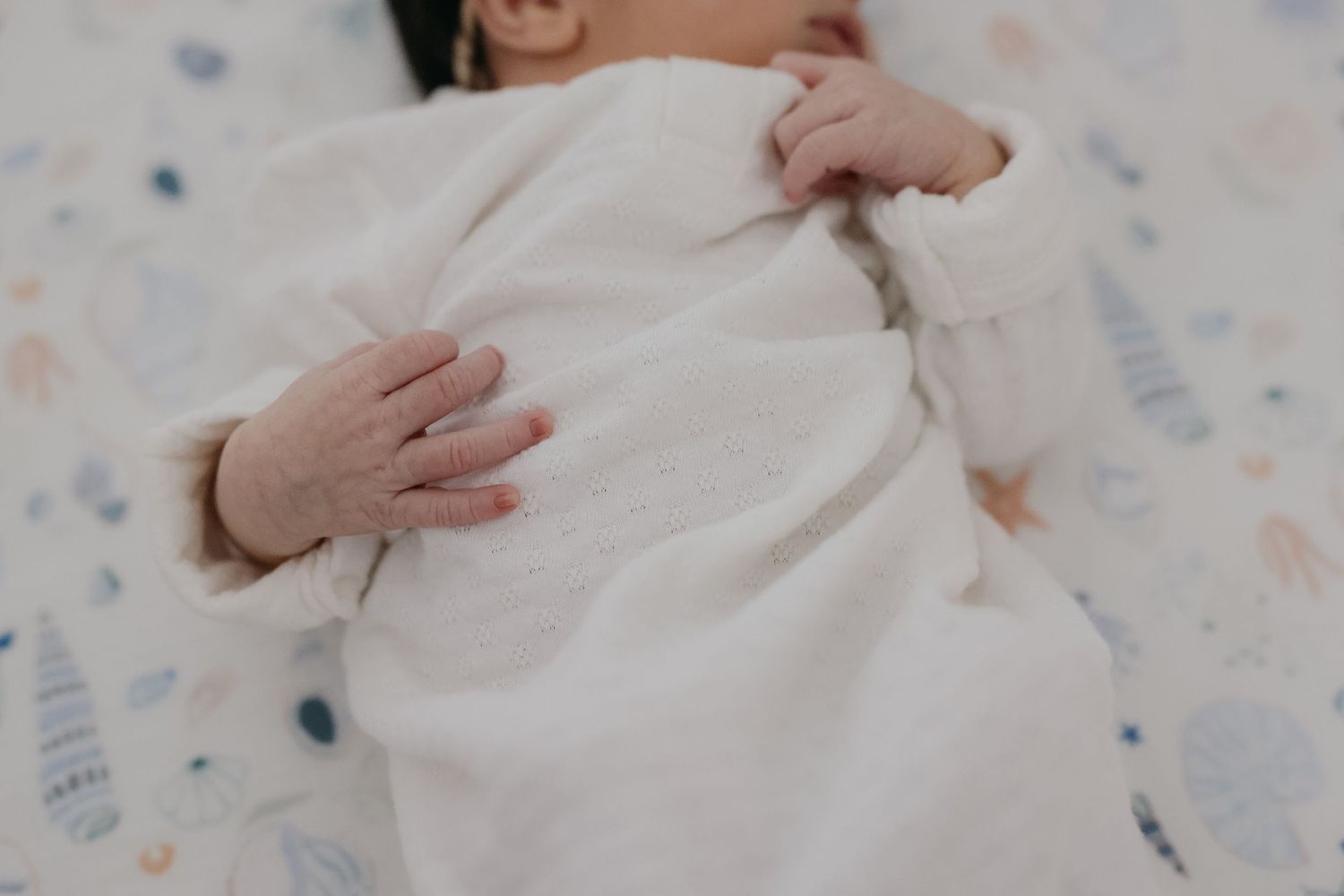 Close-up of gentle hands resting on a baby wearing white against a patterned background.
