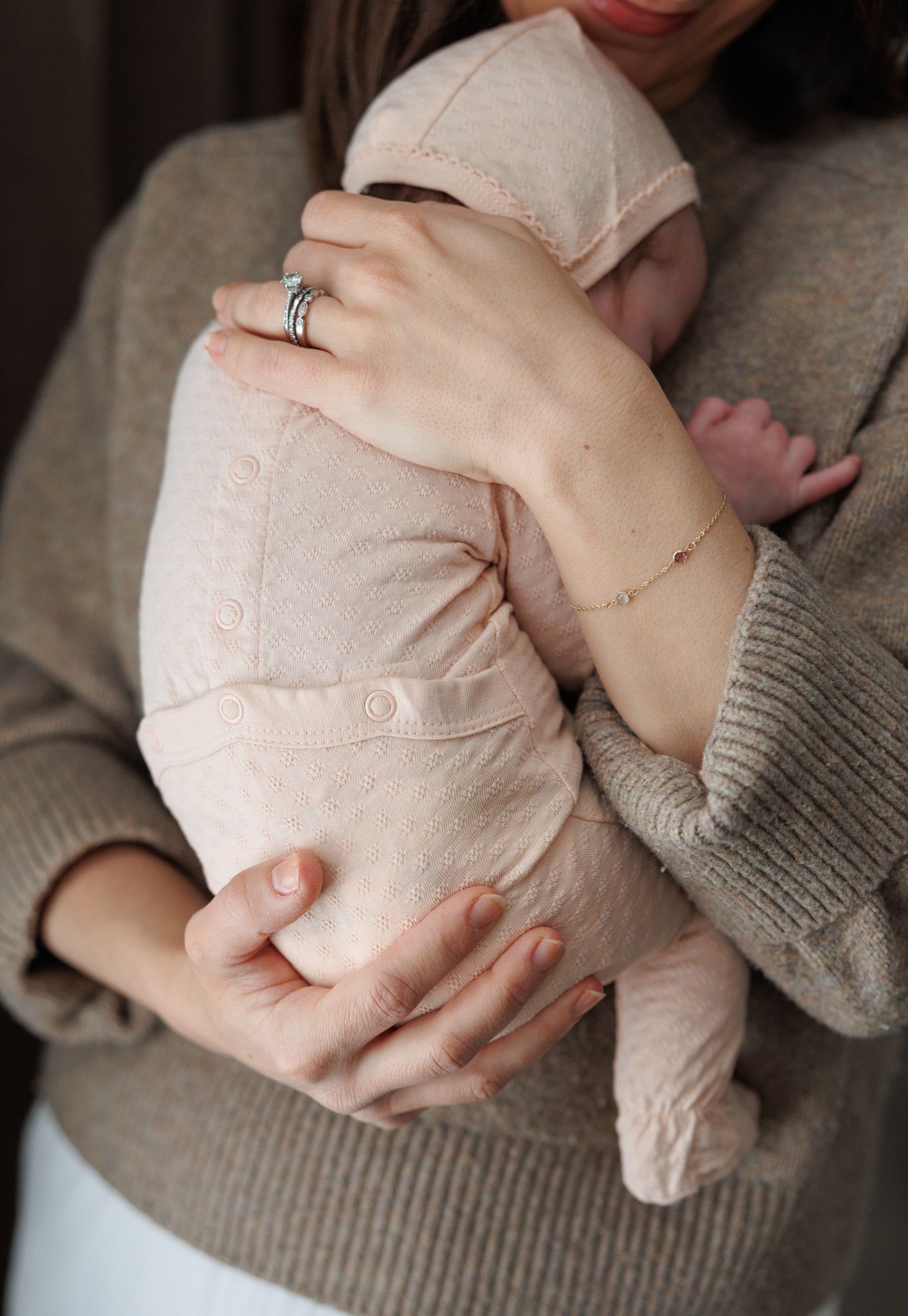 Close-up of a mother in a gray knit sweater cradling a newborn in a soft pink onesie, showcasing delicate jewelry.