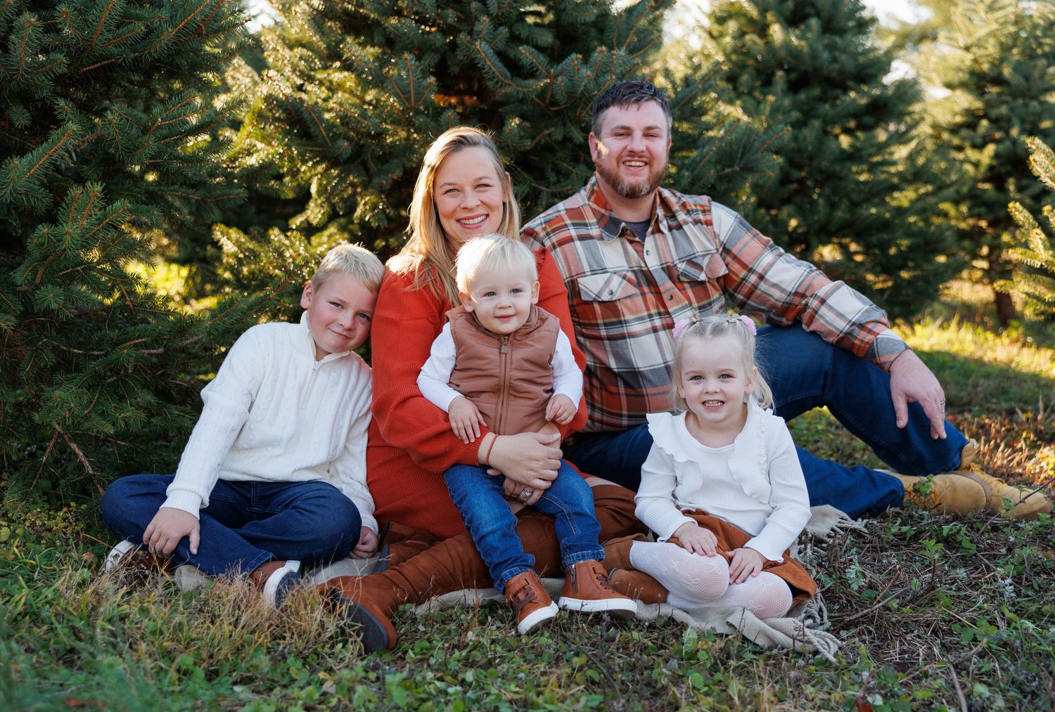 A family sits together outdoors on grass with evergreen trees in the background during a fall photo session.