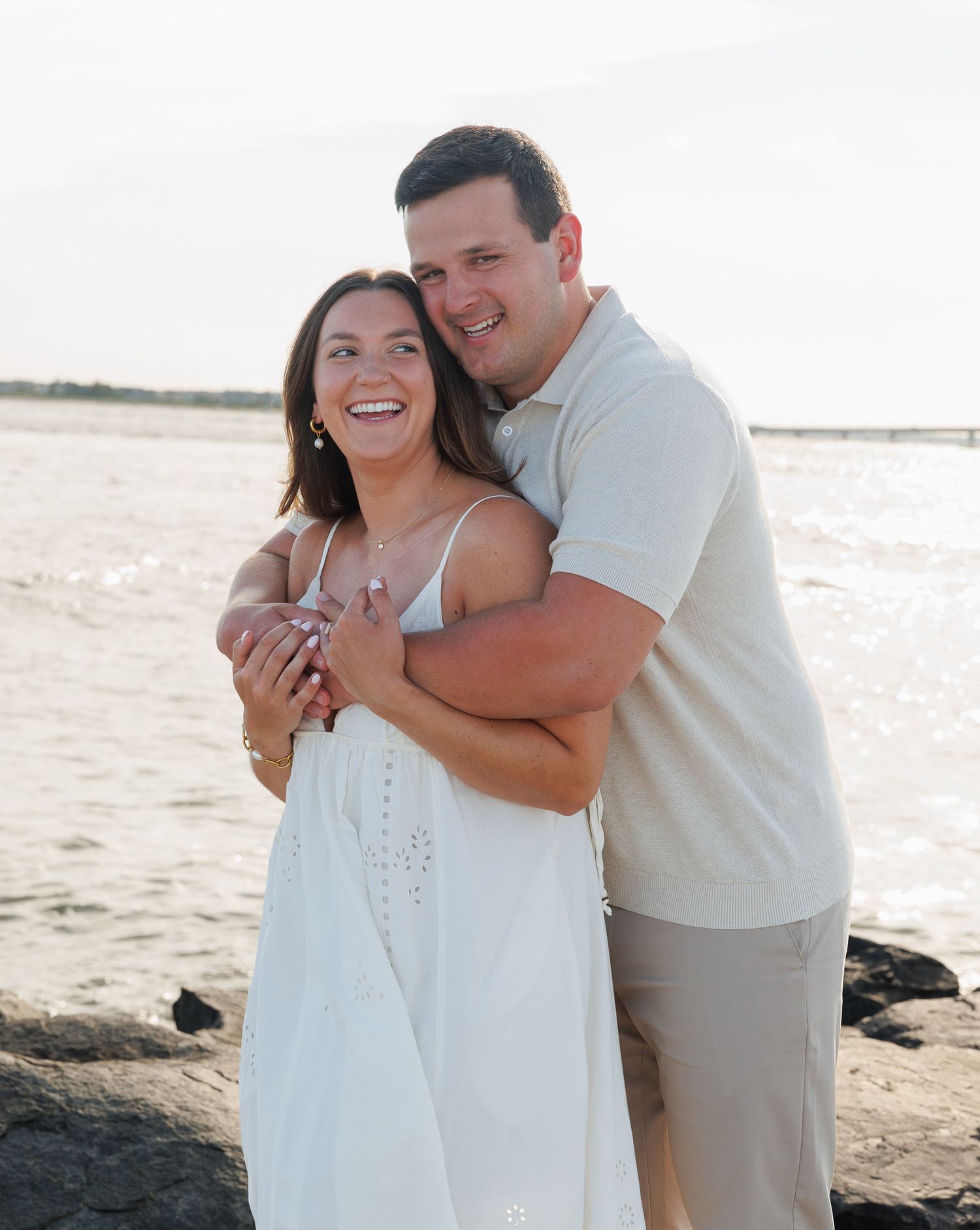 A smiling couple embraces on a rocky beach at sunset, woman in white dress, man in beige polo and khaki shorts.