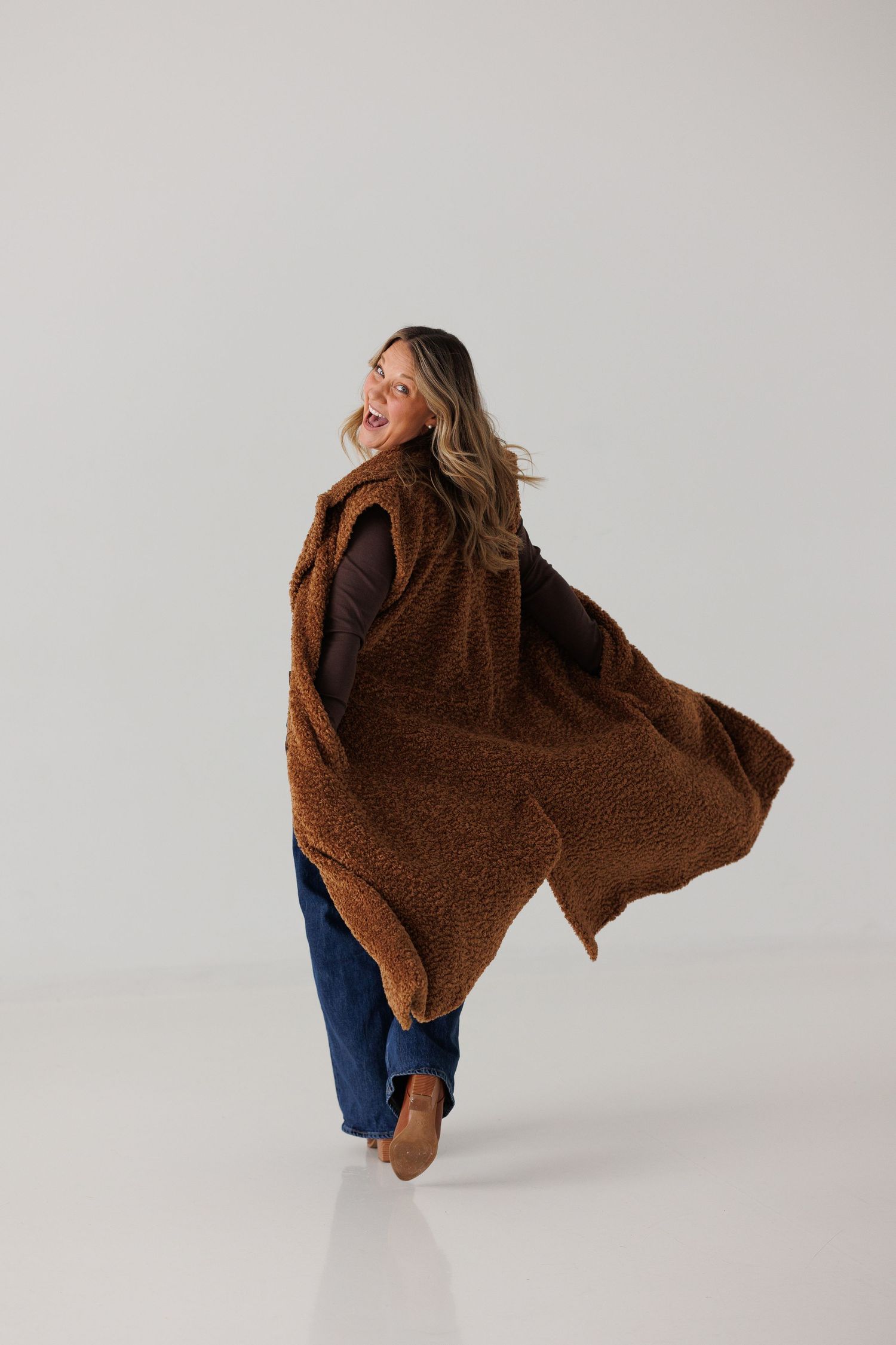 Woman twirling in a brown teddy fleece vest over dark jeans and brown boots against a white background.