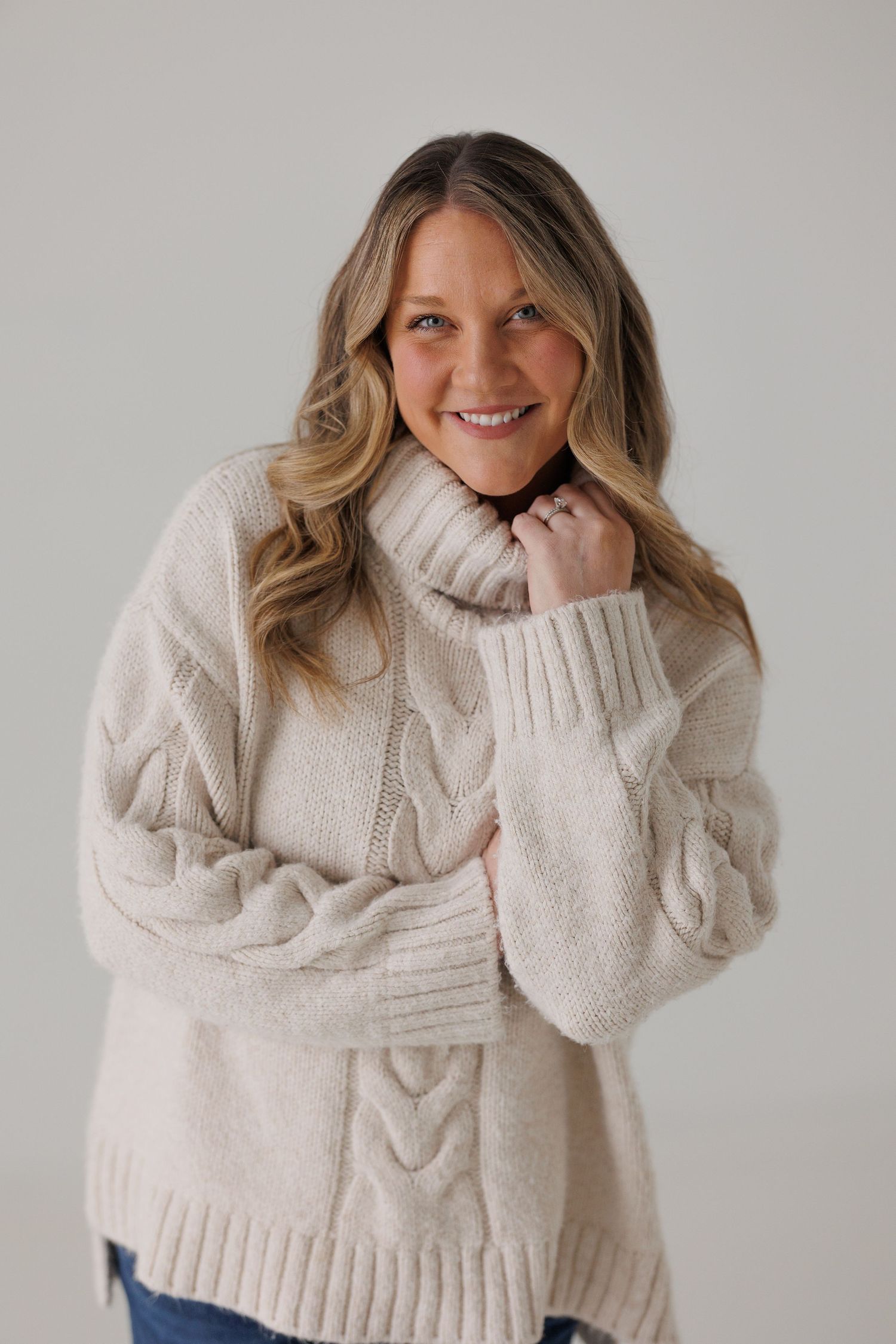 A cozy cream cable knit turtleneck sweater styled with blue jeans in a studio setting.