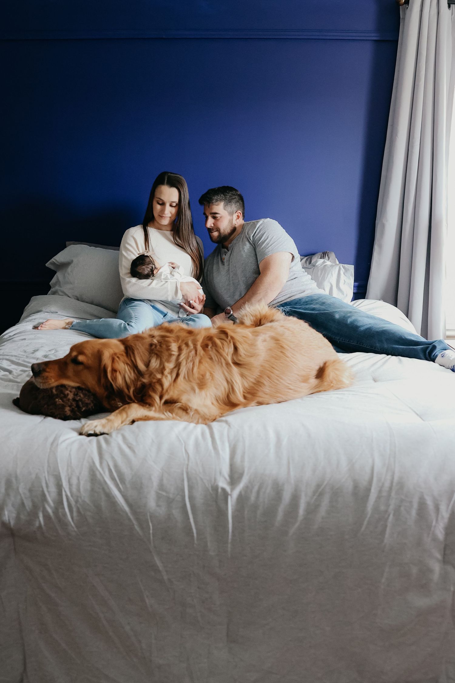 A couple rests on a bed with their newborn baby and golden retriever dog.