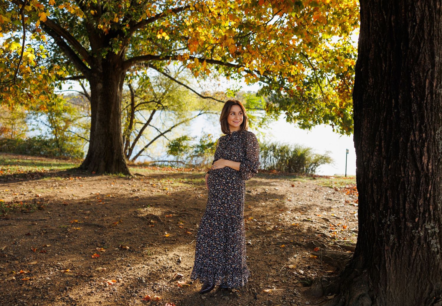 Person in a floral dress stands among autumn trees with golden leaves during a fall photo session.