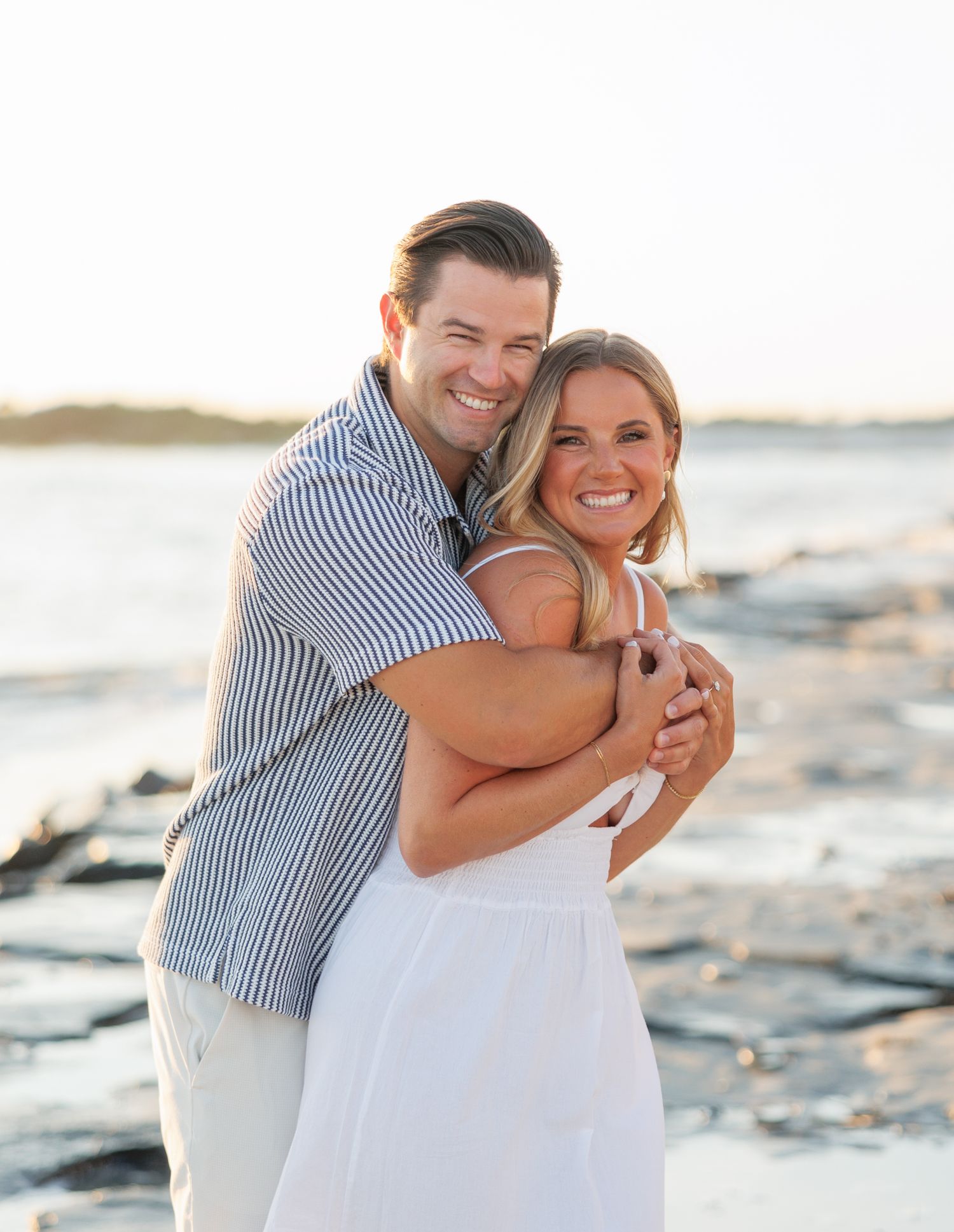 A couple embraces and laughs together during a sunset photoshoot on a rocky beach.
