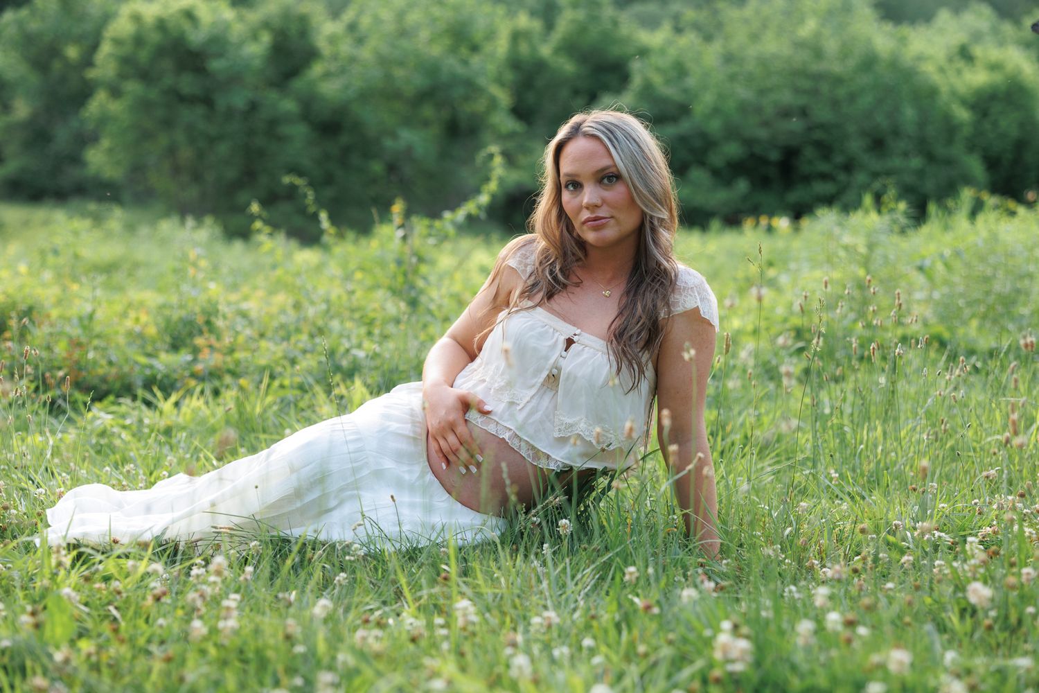 Pregnant woman in white lace dress sitting in green meadow with wildflowers, outdoor maternity photography.
