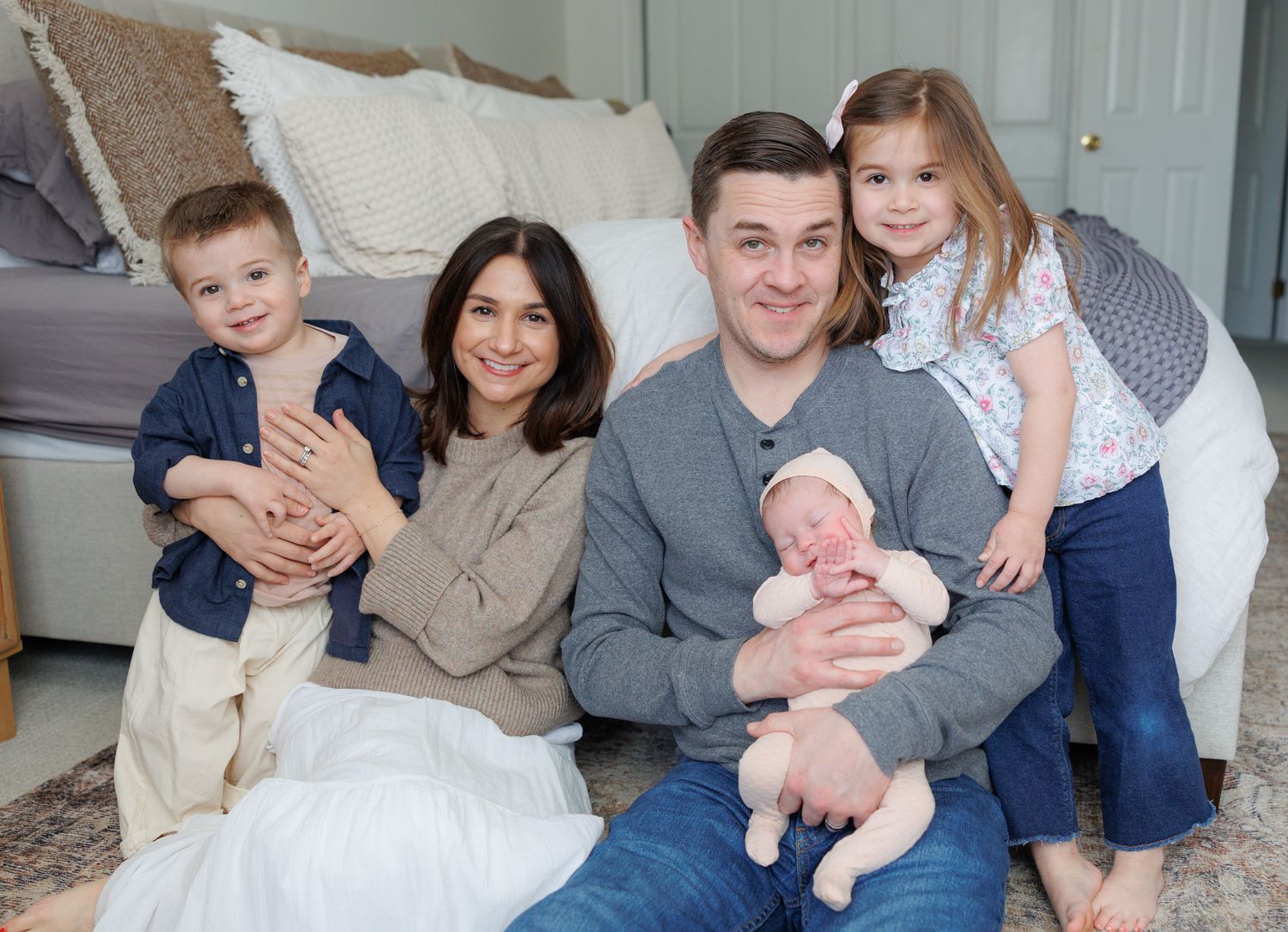Happy family of five posing together, parents holding three young children including a newborn baby, sitting in a cozy bedroom.
