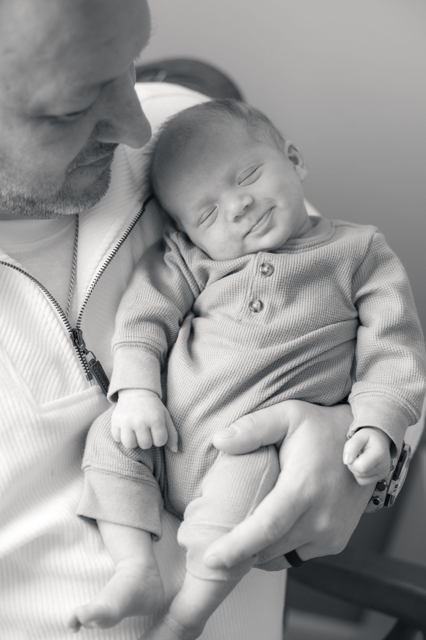 Black and white photo of a father cradling his sleeping newborn baby dressed in a ribbed onesie.