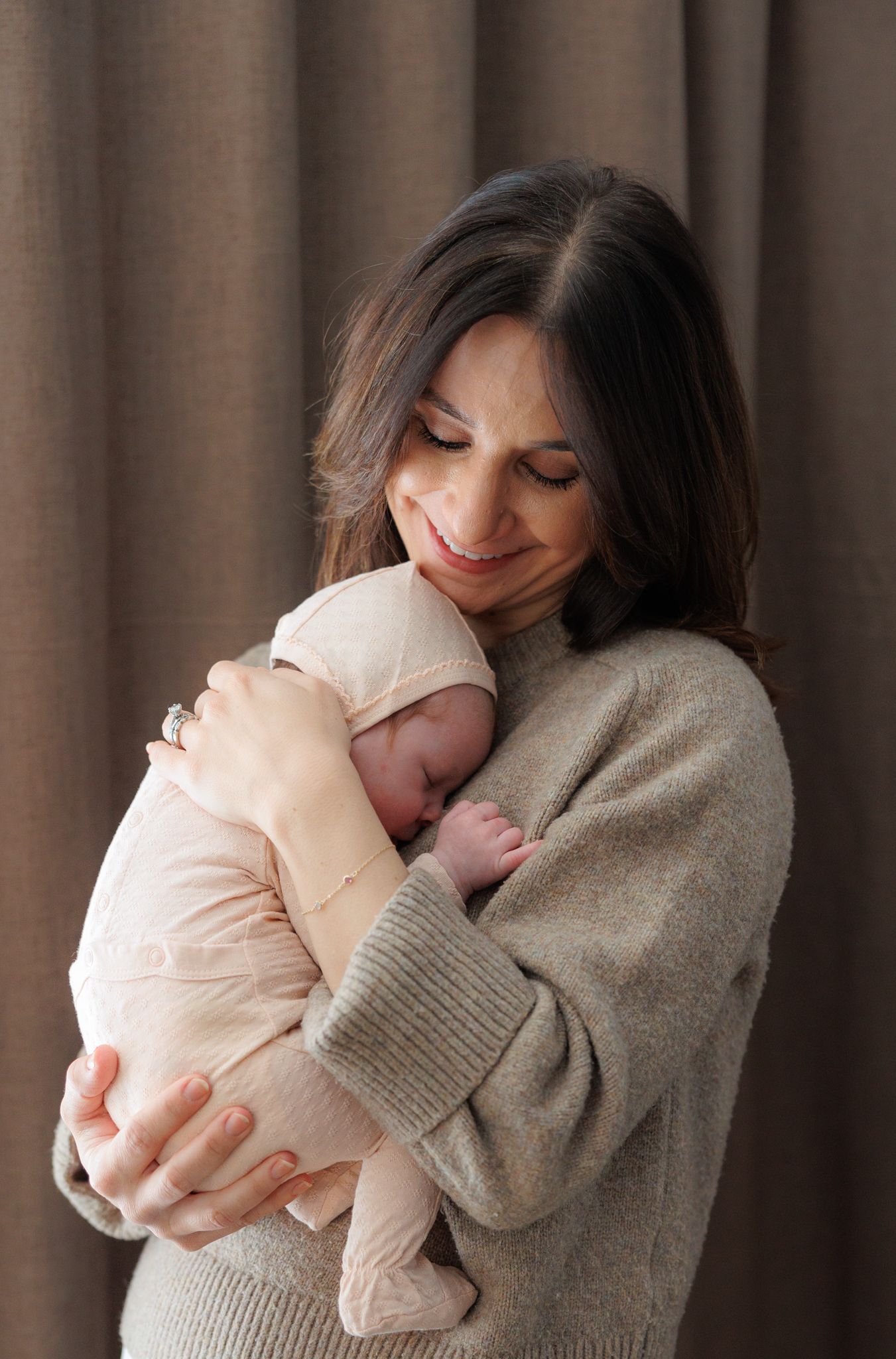 A mother tenderly holds her newborn baby, smiling softly, wearing a beige sweater against a neutral curtain backdrop.