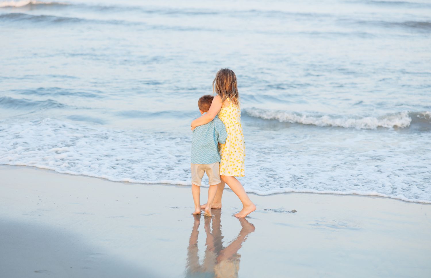 Two figures in beachwear holding hands while walking along the shoreline as gentle waves roll in at dusk. Longport, NJ