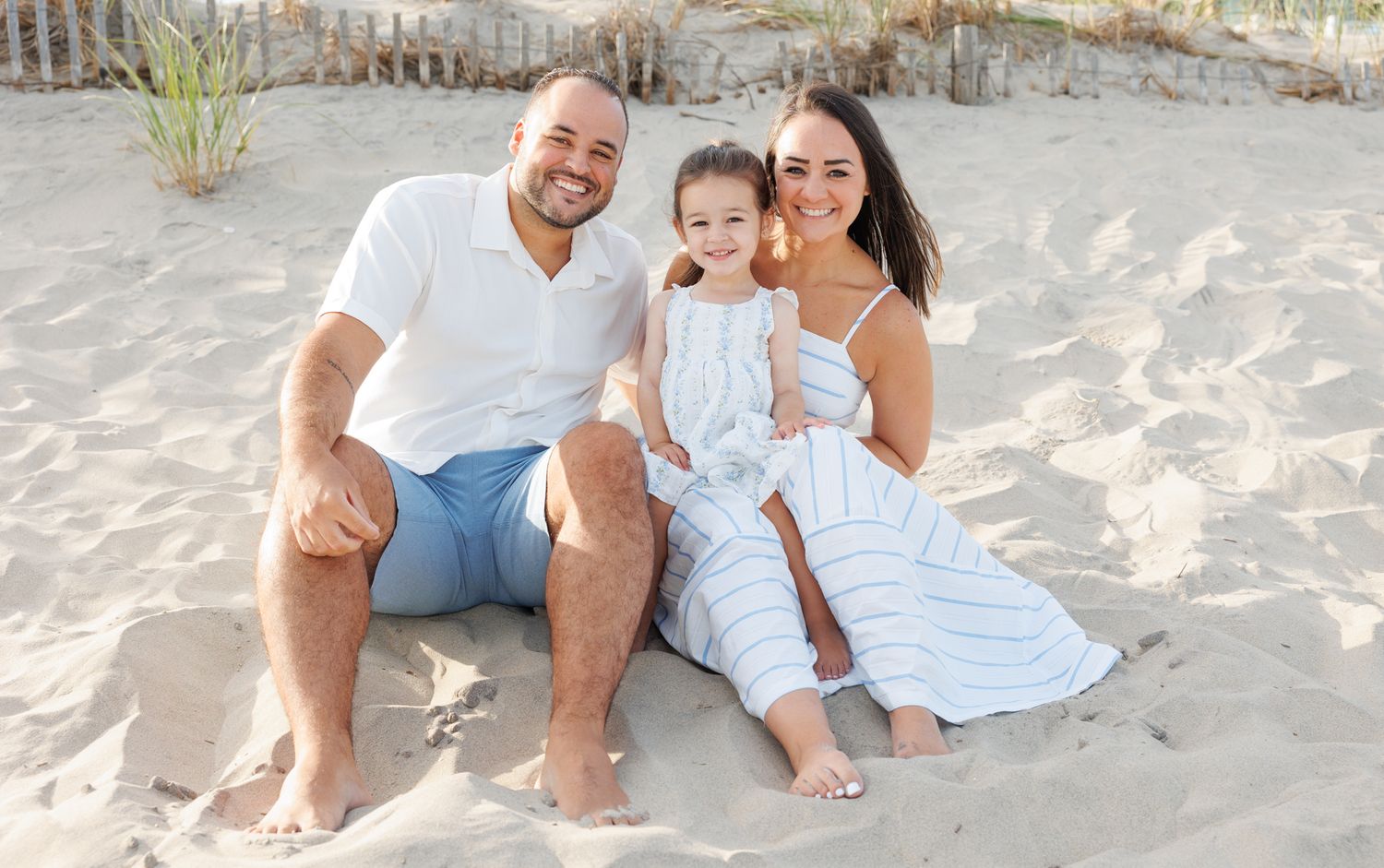 Family of three smiling on sandy beach, parents sitting with young daughter in white outfits on a sunny day.