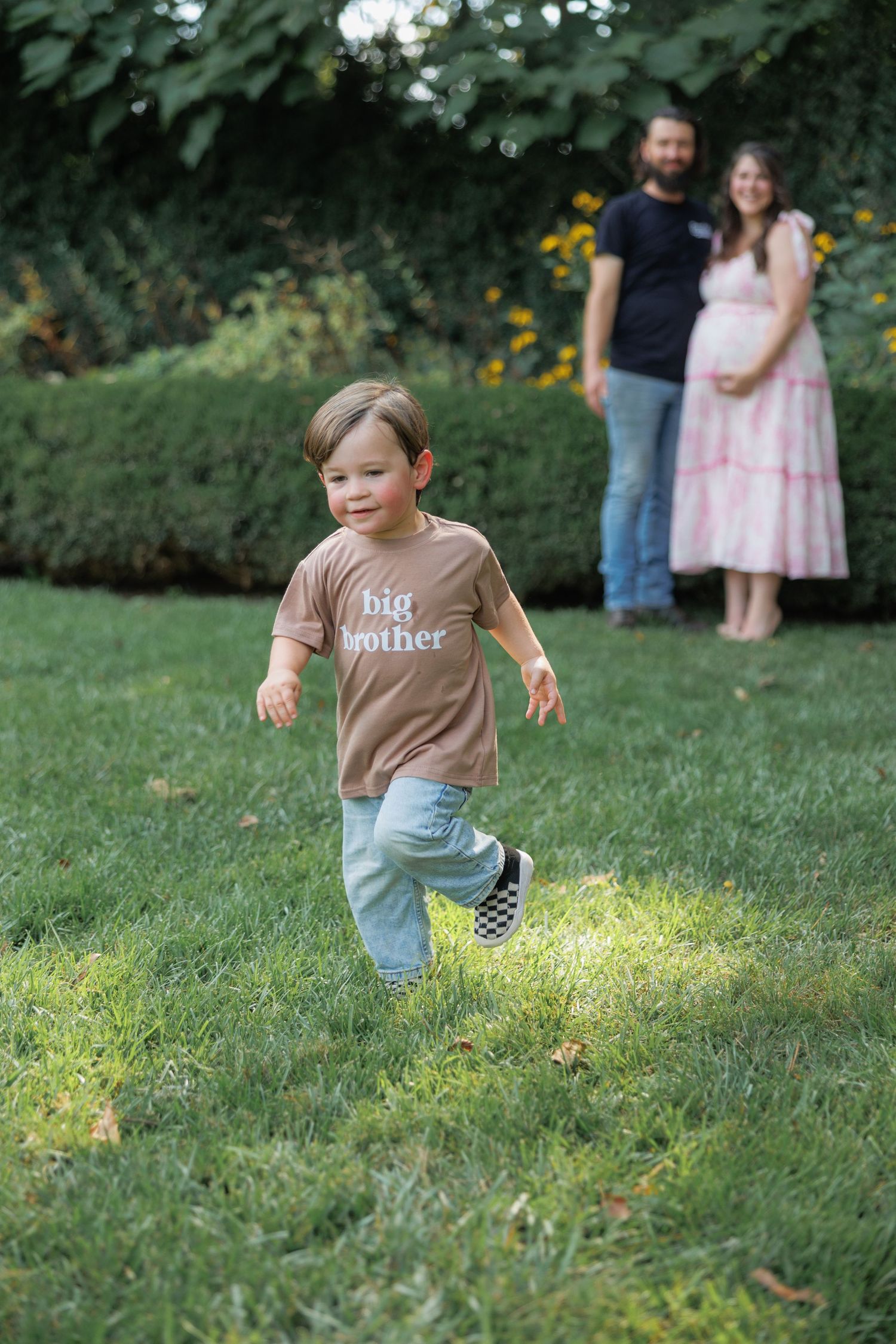 A small child wearing a brown shirt and jeans runs playfully through a grassy area on a sunny day.