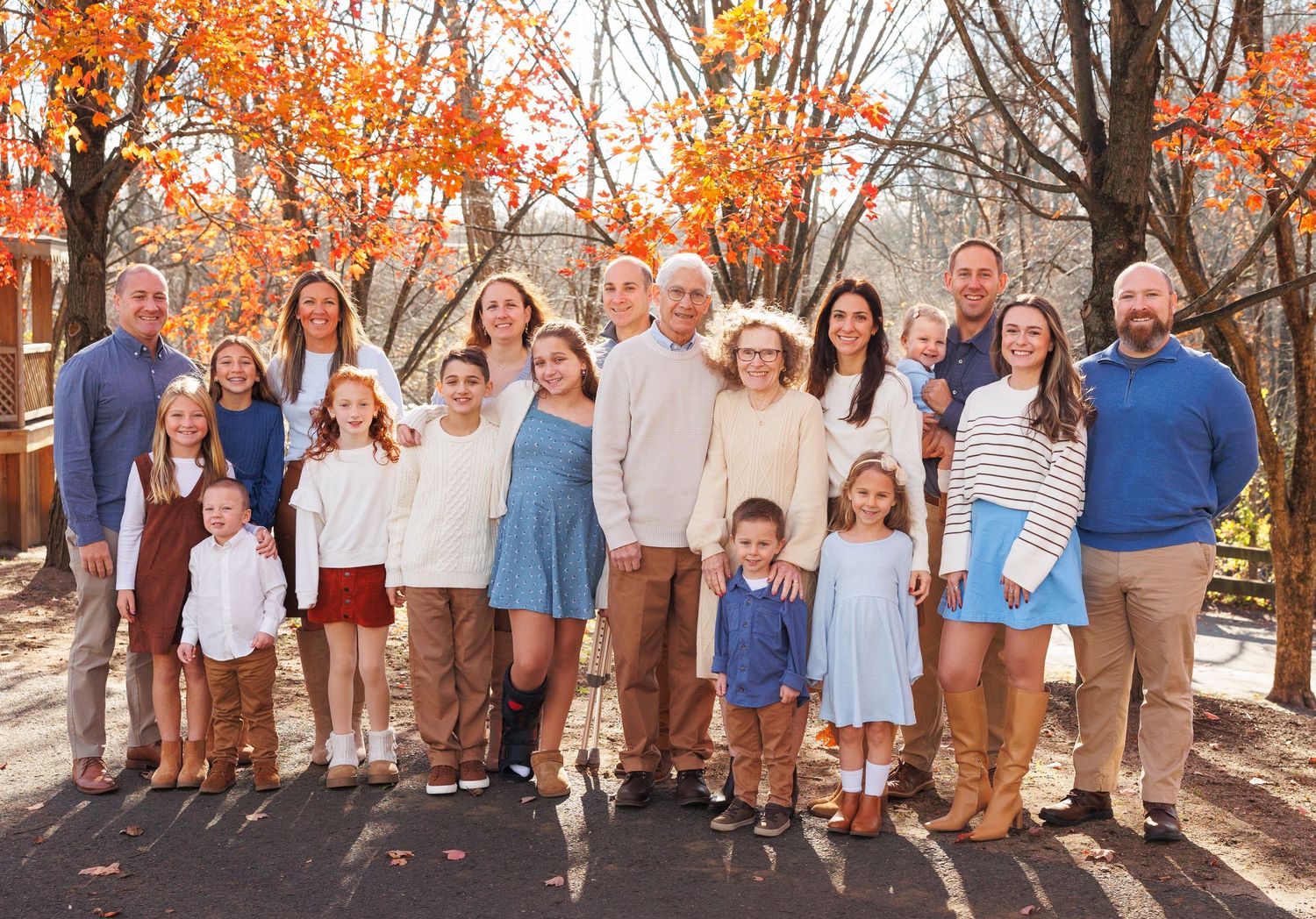 A large group stands together outdoors during autumn with orange fall foliage visible in the trees behind them.