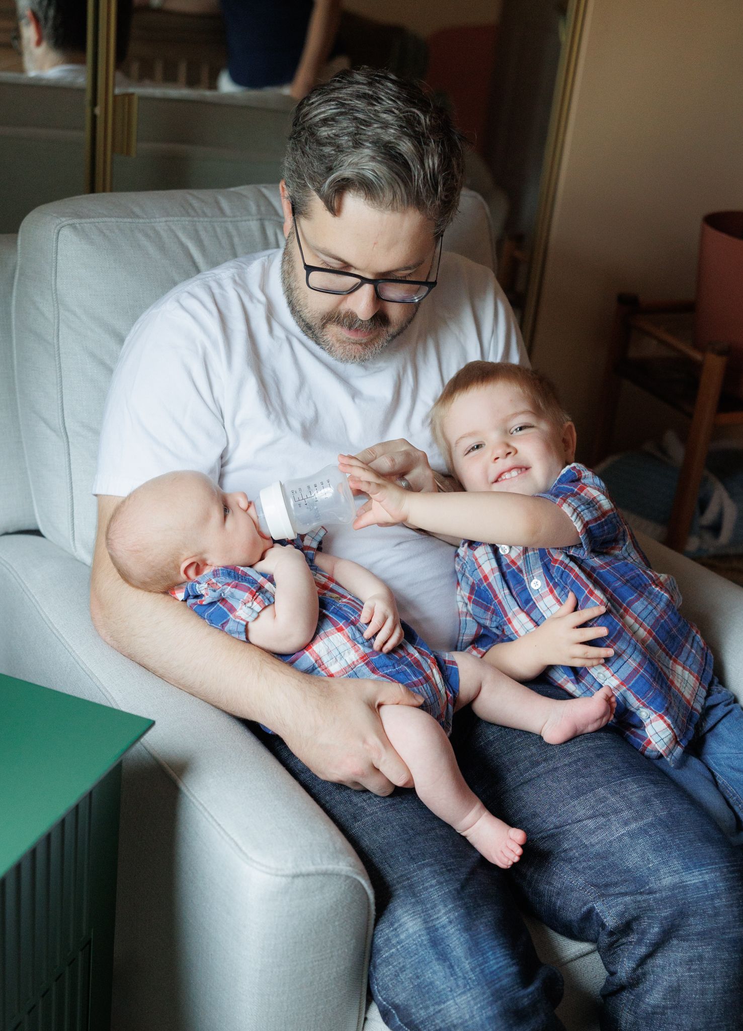Father in glasses sits in armchair bottle-feeding newborn while toddler in plaid shirt smiles beside him.