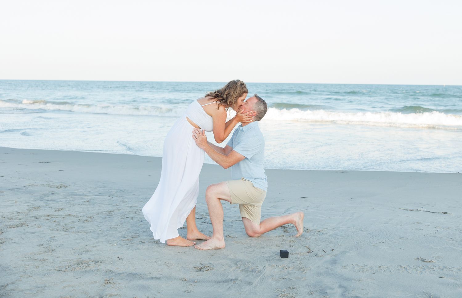 Couple in white outfits shares romantic moments during a coastal beach photoshoot at sunset.