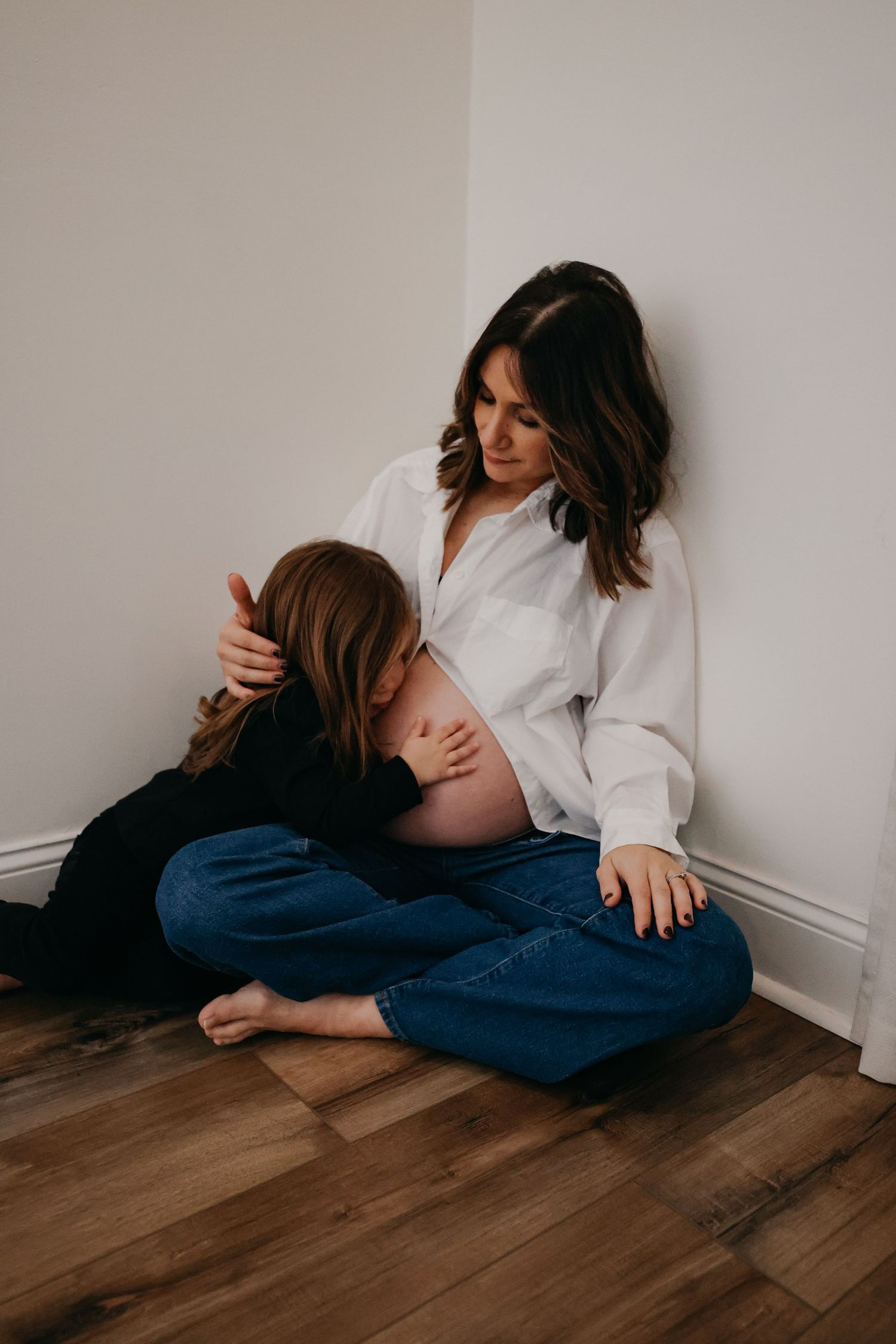 A pregnant woman in a white button-down shirt and jeans sits on hardwood floor with her child.