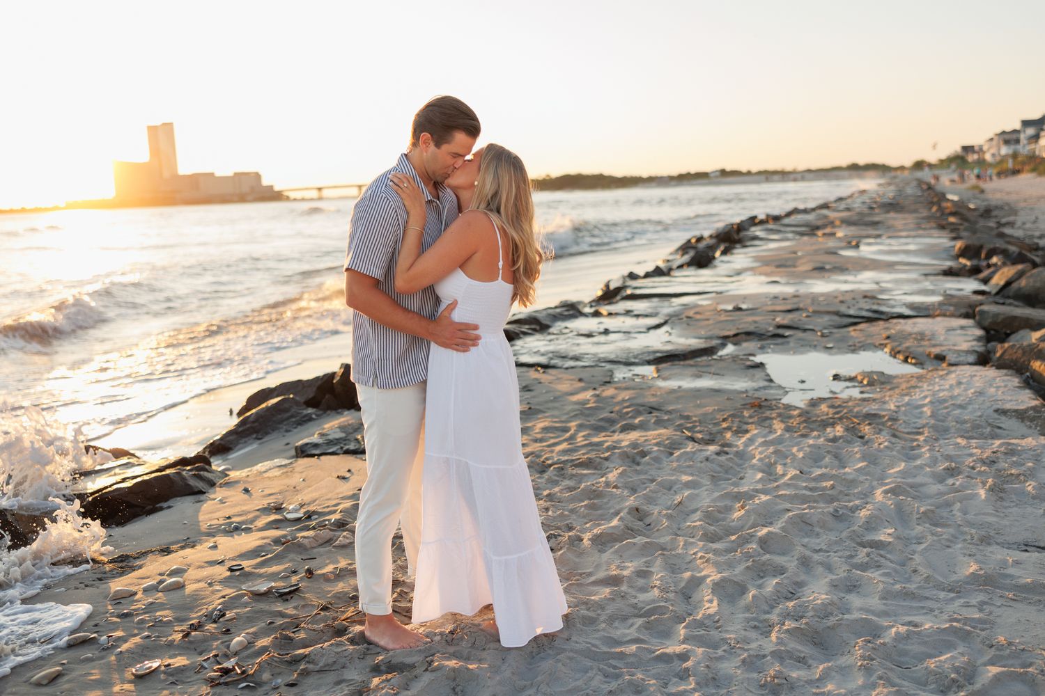 A couple shares a romantic moment on rocky coastal shores at sunset, wearing light colored outfits.