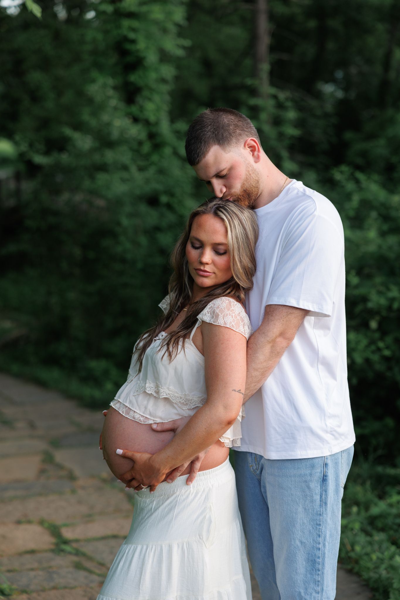 Couple sharing a tender maternity moment outdoors, man kissing woman's head while cradling her baby bump on a garden path.
