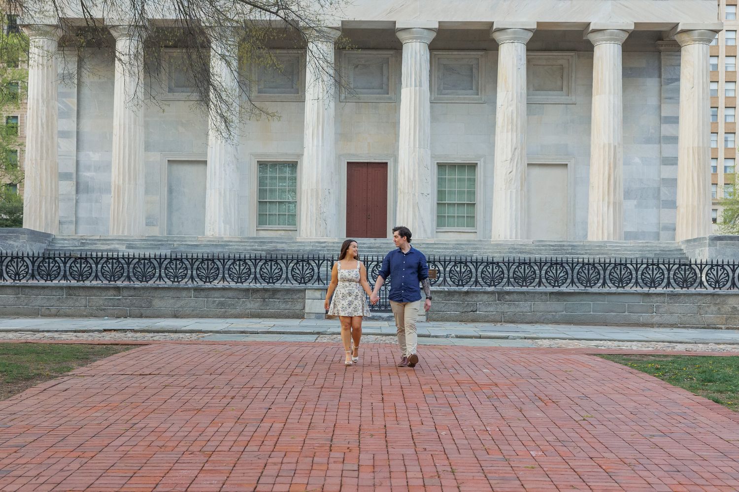 Person walks along red brick pathway beside stone pillars and white building exterior.