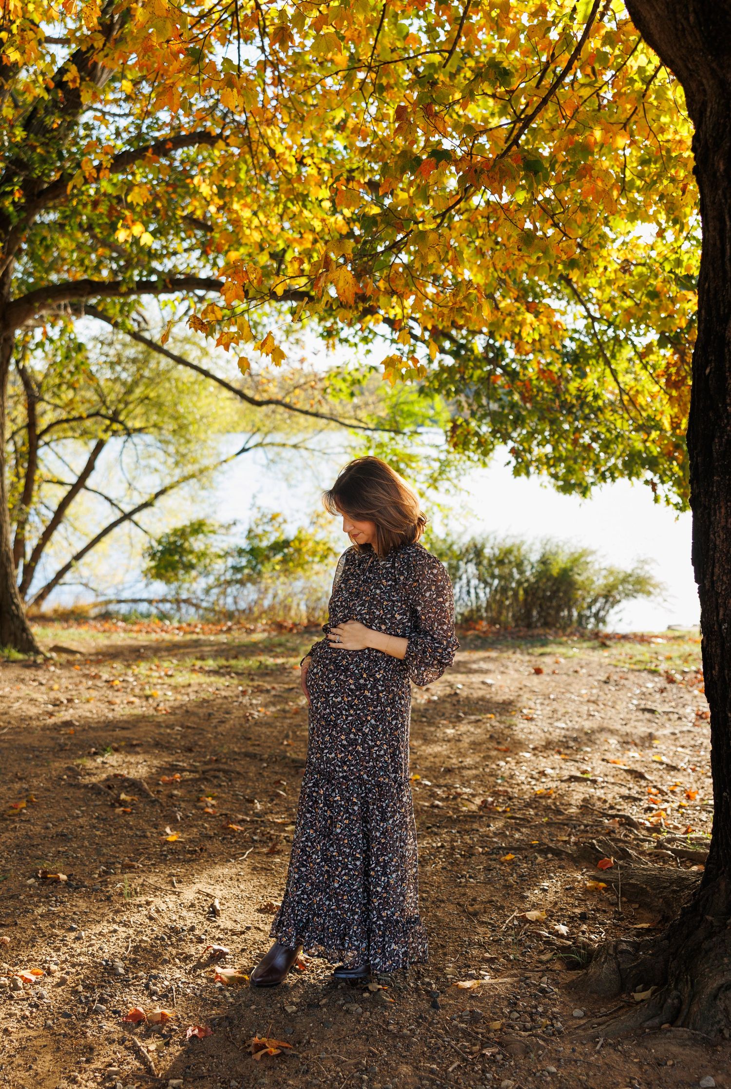A person in a polka dot dress stands beneath golden autumn trees near a lake on a sunny fall afternoon.