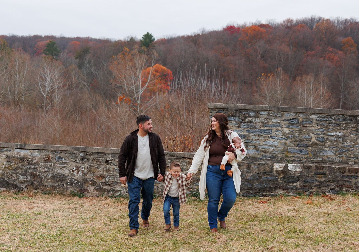 A family walks together on an autumn day near a stone wall with fall foliage on the hillside in the background.