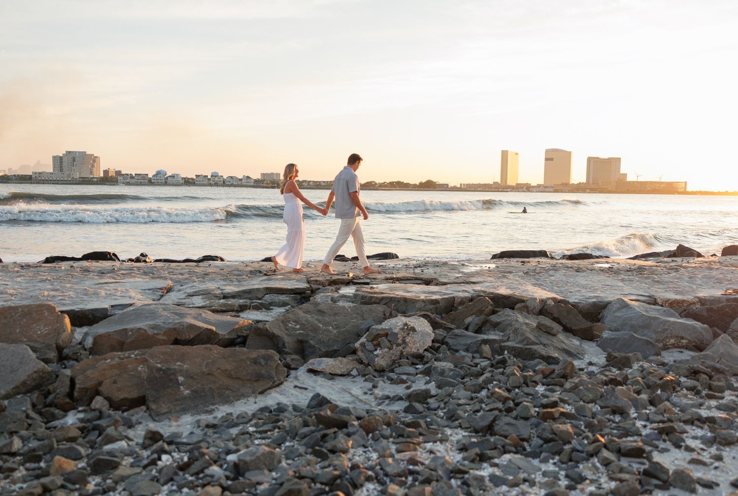 Couple walking along rocky coastal shoreline during golden sunset with cityscape in background.