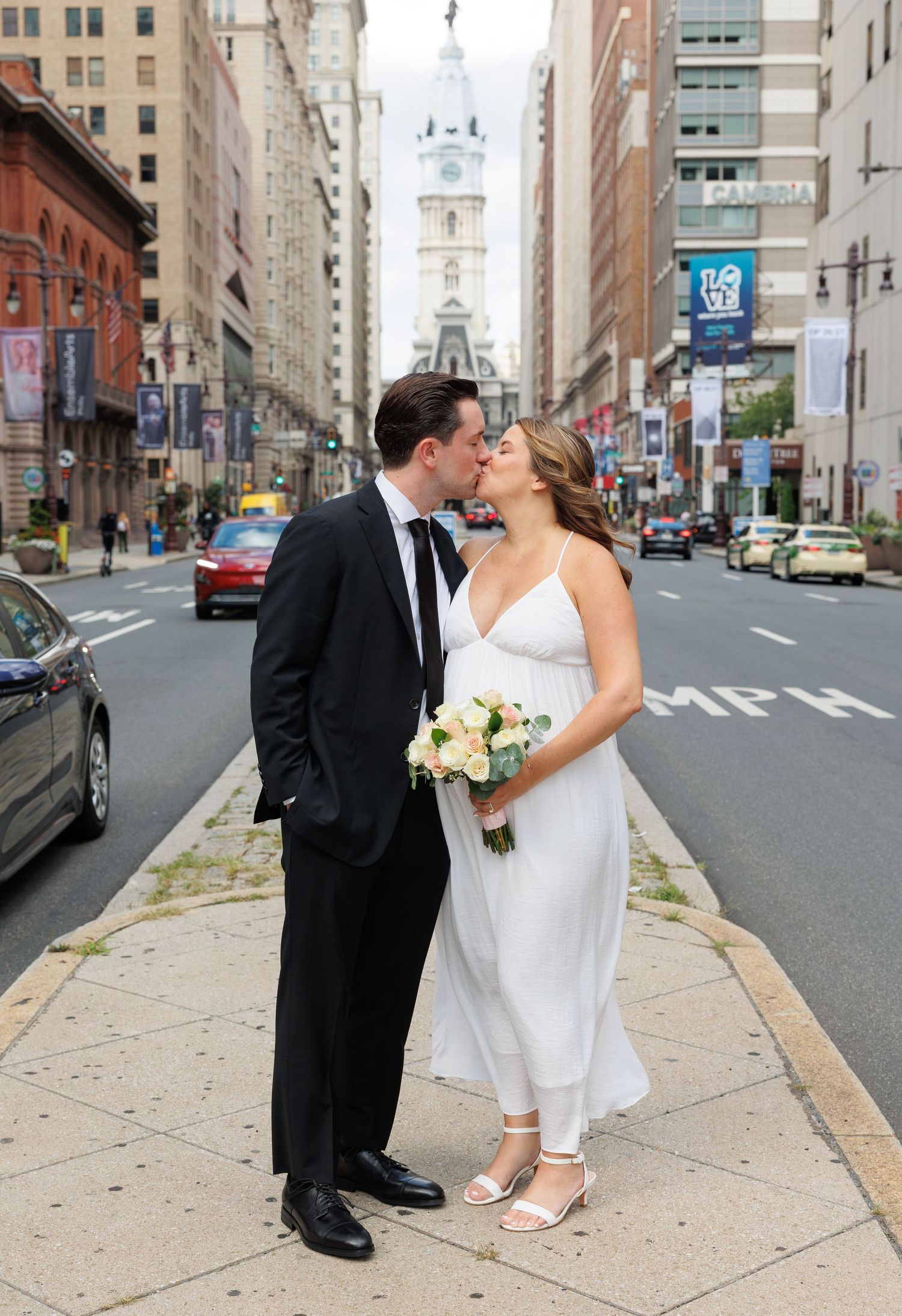 Newlyweds kissing on a Philadelphia street corner in wedding attire, with City Hall tower visible in the background.